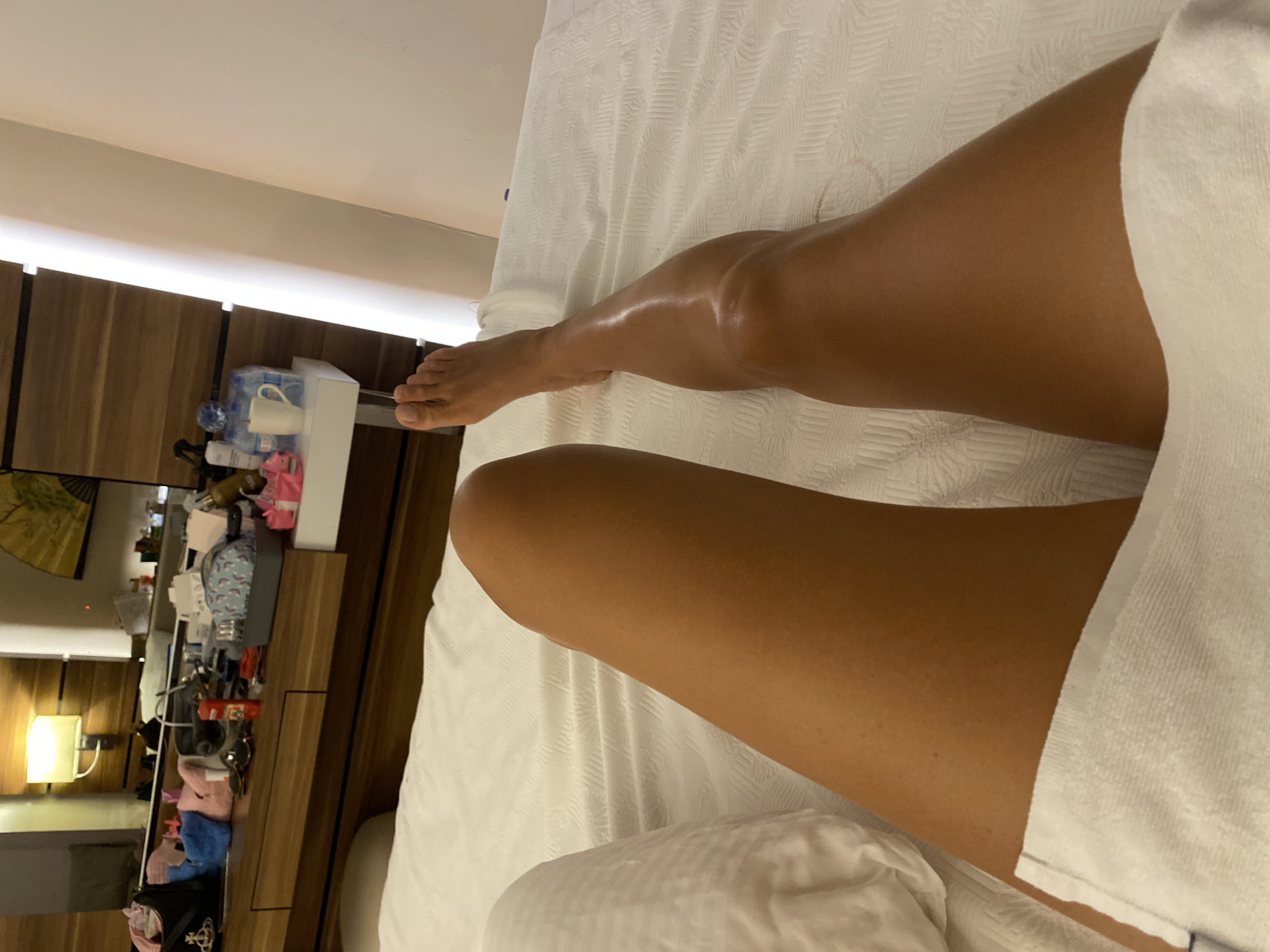 The image shows a person's legs, from the thigh down to the foot, resting on a white, textured surface that appears to be a bed or massage table. The legs are tanned and appear to be freshly moisturized, with a visible sheen on the skin of the lower leg. A white towel is draped over the lower thigh of the left leg. In the background, a dresser or vanity is visible, cluttered with various items including bags, bottles, and a mirror. A lamp is mounted on the wall above the dresser. The overall setting suggests an indoor environment, possibly a hotel room or spa. The location is not specified in Aksu, Turkey in this image.