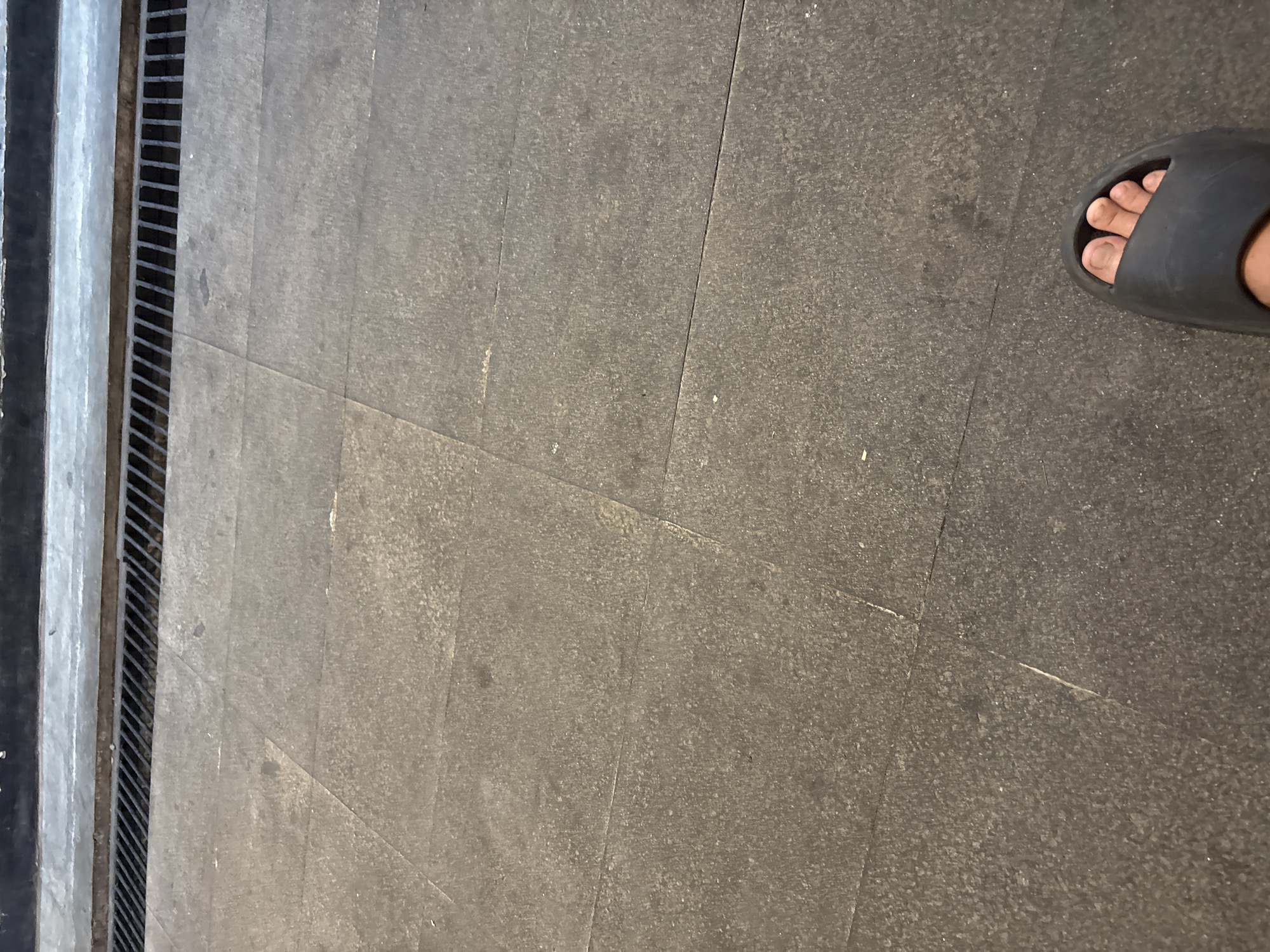 The image shows a person's bare foot wearing a black slide sandal, positioned on a tiled floor. The tiles are large, square, and dark gray with a speckled texture. A metal grate, likely a drain, runs horizontally across the top portion of the image, adjacent to a dark-colored wall or curb. The scene appears to be outdoors, possibly on a sidewalk or plaza in Cirebon, Indonesia, given the presence of the drain and tiled public space often found in urban environments. The lighting suggests it is daytime, with some shadow cast by the grate.