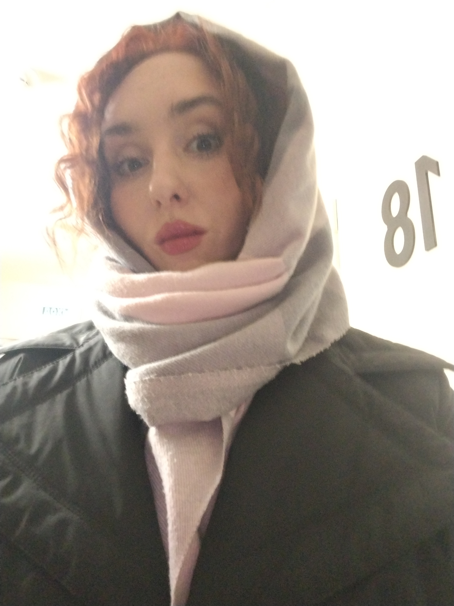 A woman with red curly hair is wearing a light pink and gray scarf draped over her head and around her neck. She is wearing a dark-colored jacket or coat. The background is blurred, but the number "81" is visible on a white surface. There is no indication of Izhevsk, Russia, in the image.