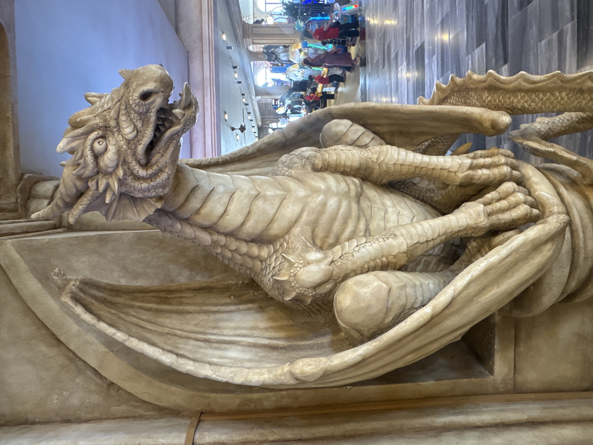 The image features a large, intricately detailed statue of a dragon made of a light, earthy-toned material, possibly stone or resin, positioned prominently in the foreground. The dragon is depicted in a dynamic, possibly roaring or startled pose, with its head uplifted and facing towards the left of the frame. Its mouth is agape, revealing fangs, and its eyes are wide open, conveying a fierce expression. The sculpture showcases detailed scales, spiky protrusions along its back, and large, textured wings folded close to its body. Muscular limbs with clawed hands are also clearly visible.
In the background, a brightly lit indoor environment extends, resembling a long corridor or hall within an entertainment complex. Several people are visible further down this hall, engaged in playing various brightly lit arcade machines, indicating a leisure or gaming setting. The floor of the hall has a patterned design, and the walls exhibit architectural details.
The scene suggests an active, indoor recreational space, likely bustling with visitors. The time of day is not evident, as artificial lighting illuminates the entire area. The dragon statue's dramatic expression and impressive scale are notable details, serving as a striking focal point within the venue. No visible text is present in the image. The location is an entertainment venue in Đà Nẵng, Vietnam.