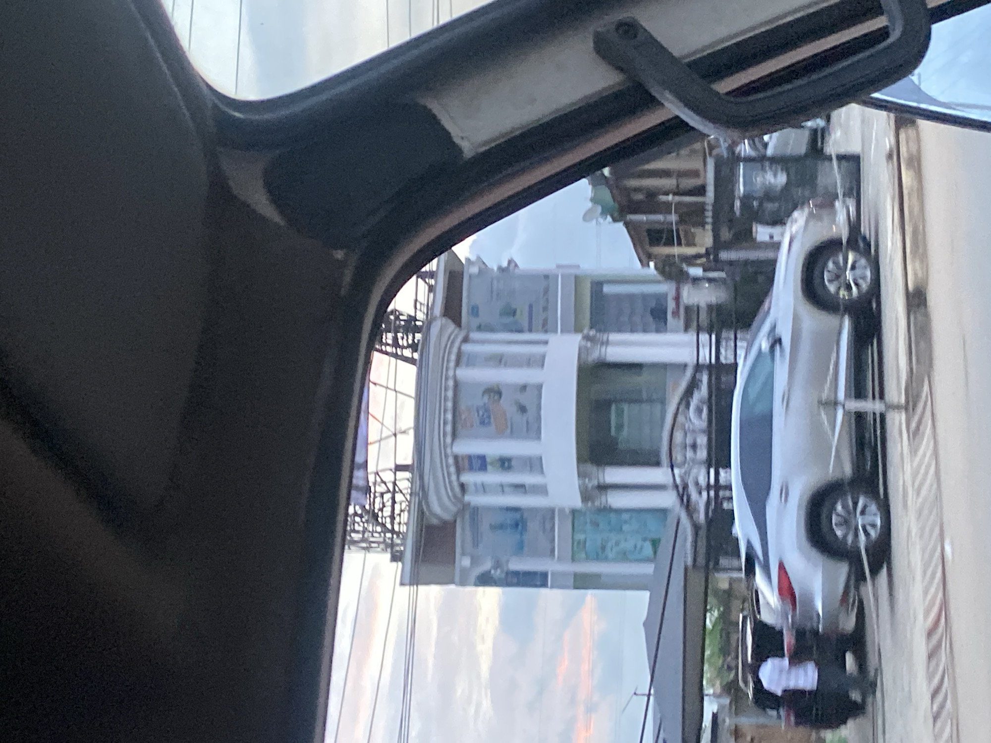 The image, taken from inside a vehicle, shows a street scene in Ikorodu, Nigeria. A multi-story building with a distinctive curved facade and white columns is prominently featured. The building appears to be a commercial establishment, with signage visible on its exterior. In the foreground, a silver SUV is parked on the street, and a person in a white and striped shirt is walking near a dark vehicle. The sky is partly cloudy with hints of pink and orange, suggesting it may be dusk or dawn. The overall environment appears to be an urban or semi-urban setting with overhead wires and other structures indicative of a populated area.