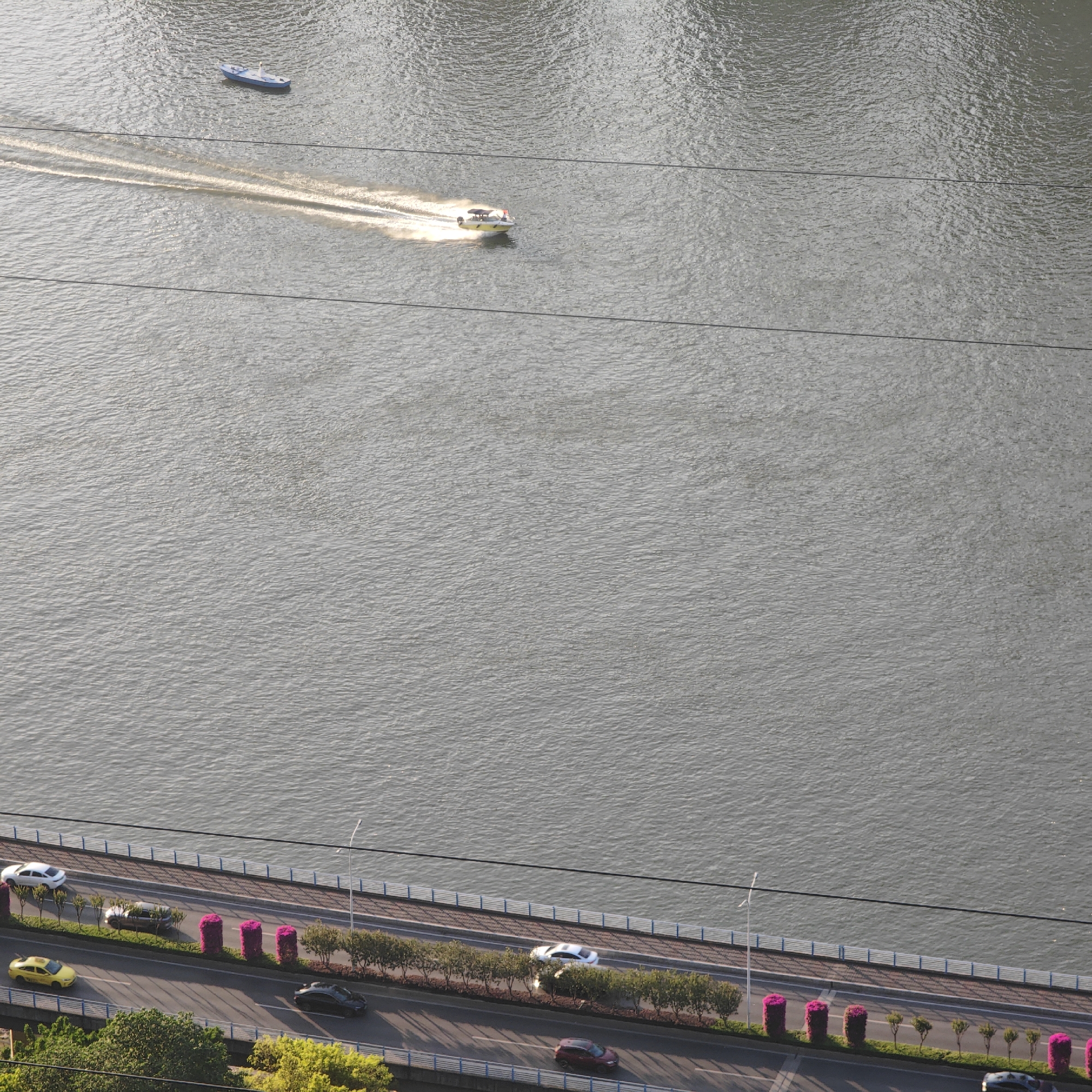 The image presents an elevated view of a waterfront area in Yuzhong District, China. A large body of water dominates the upper portion of the frame, with two boats visible; one smaller boat is near the top left corner, and a larger, yellow boat is traveling across the water, leaving a visible wake. Three parallel electrical lines stretch horizontally across the water.
Along the bottom of the frame, a road runs alongside the water. A pedestrian walkway, bordered by a railing, separates the road from the water. Several vehicles are on the road, including a yellow taxi, white sedan, and dark-colored car. Flowerbeds and landscaping are arranged along the median and sidewalk.
The scene is captured in daylight with a somewhat overcast sky, leading to soft lighting and muted colors. The angle of the photograph suggests it was taken from a high vantage point, such as a tall building.