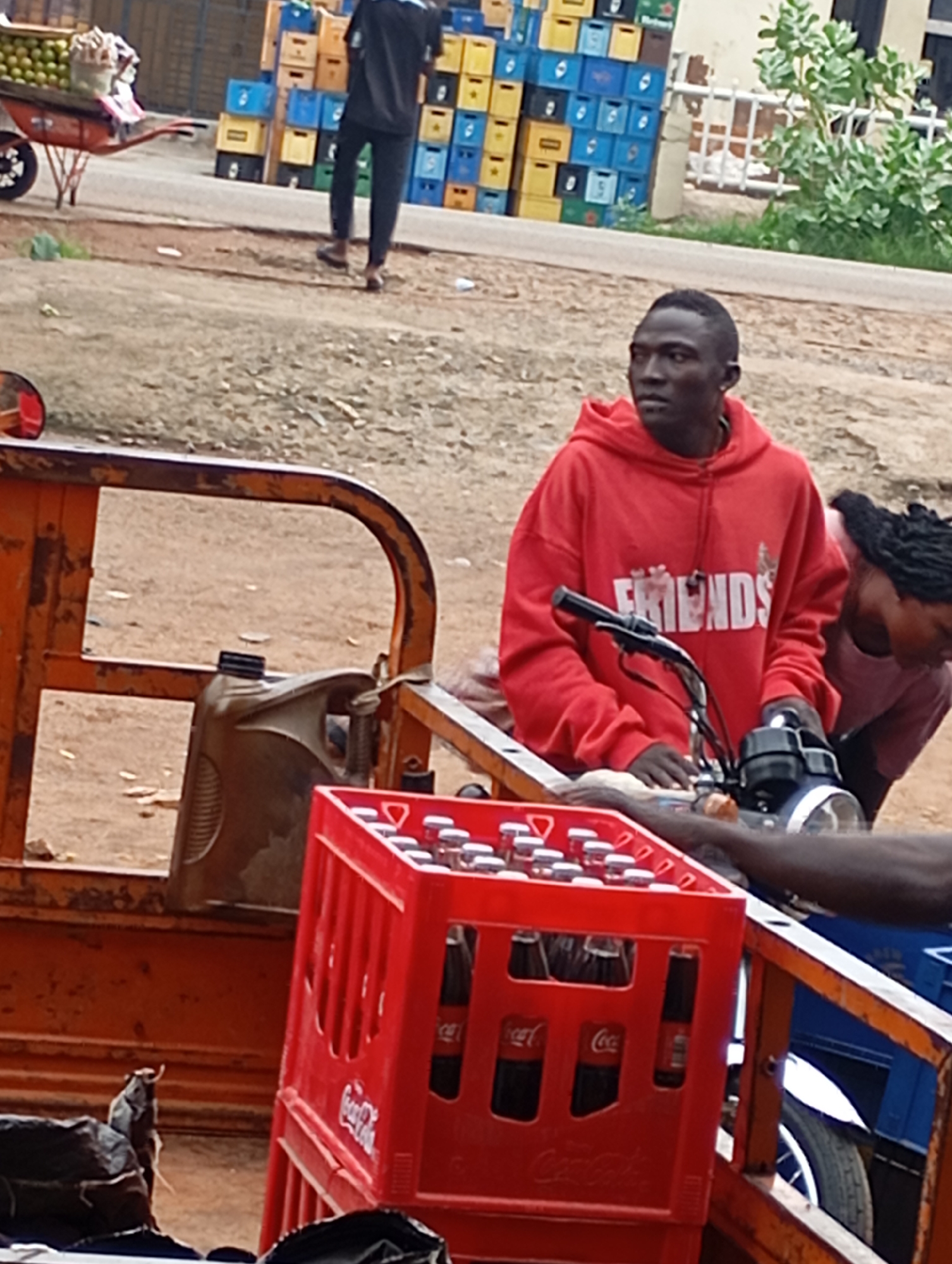 In Shendam, Nigeria, a young Black man wearing a red hoodie with "FRIENDS" printed on it is seated on a three-wheeled motorcycle, looking to his left. Behind him, a Black woman with dark braided hair, wearing a pink t-shirt, is partially visible. In the foreground, a red plastic crate filled with Coca-Cola bottles sits on the motorcycle's flatbed. The scene appears to be a street, with a dirt ground and stacks of colorful crates in the background. The overall impression is that of a commercial transaction or transportation of goods.