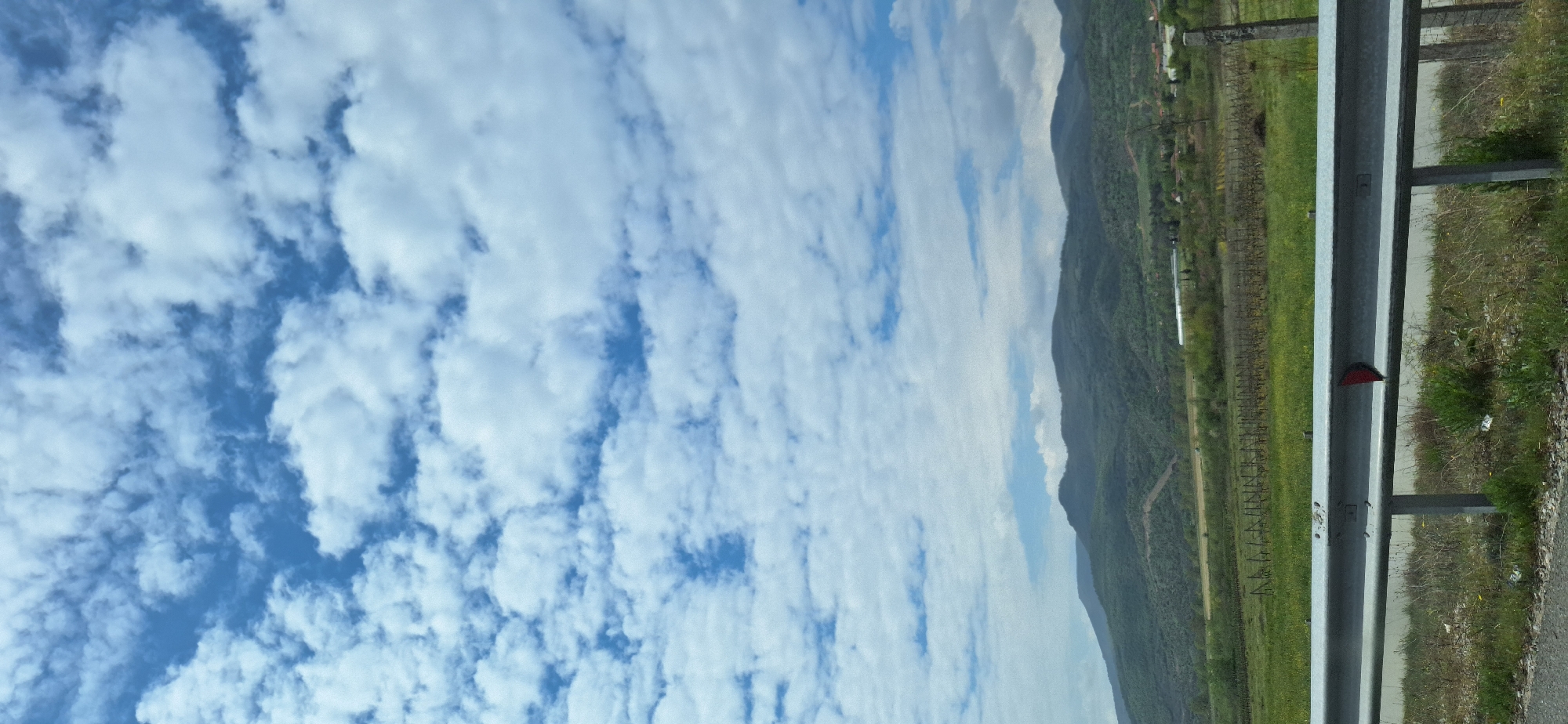The scene depicts a roadside view in what is suggested to be near Valkovo, Bulgaria. The sky is mostly blue, filled with patterned clouds. Adjacent is a mountainous landscape with greenery. At the edge of the frame are metal guardrails, with vegetation growing around them. In the distance, there appears to be a vineyard or cultivated land.