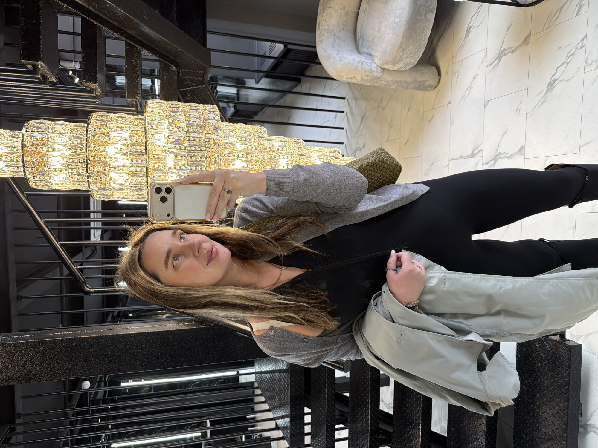 The image shows a woman standing on metal stairs. She is holding a smartphone up to a large crystal chandelier suspended above her. She is wearing black leggings, a black top, and a light gray cardigan. She also has a light gray jacket draped over her arm. Behind her is a wall with white marble tiles and a light gray chair. It appears to be a showroom. The location may be in Moscow, Russia.