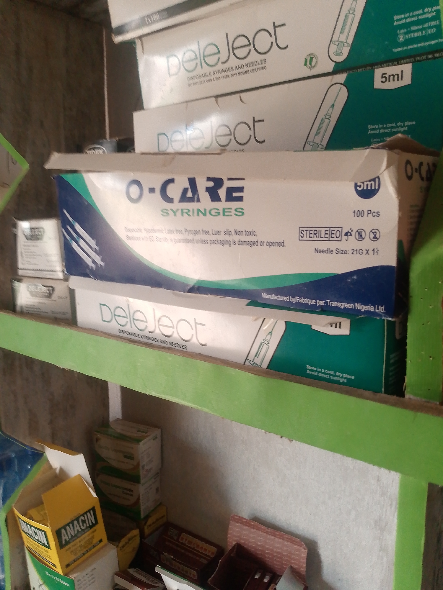 The image displays a shelf filled with medical supplies, primarily syringes and needles in their packaging. The items are stacked and arranged in a somewhat disorganized manner, suggesting a storage or retail setting. Visible brands include "Deleject" and "O-CARE SYRINGES." The packaging indicates that these are disposable and sterile products. Some boxes also mention specifications like needle size and volume. In the foreground, there are also boxes of "ANACIN" medication, suggesting this shelf holds a variety of pharmaceutical items. The environment appears to be a dimly lit indoor space, possibly a pharmacy or a medical store. There are no people visible in the image. The overall impression is one of a cluttered but functional storage area for medical supplies.