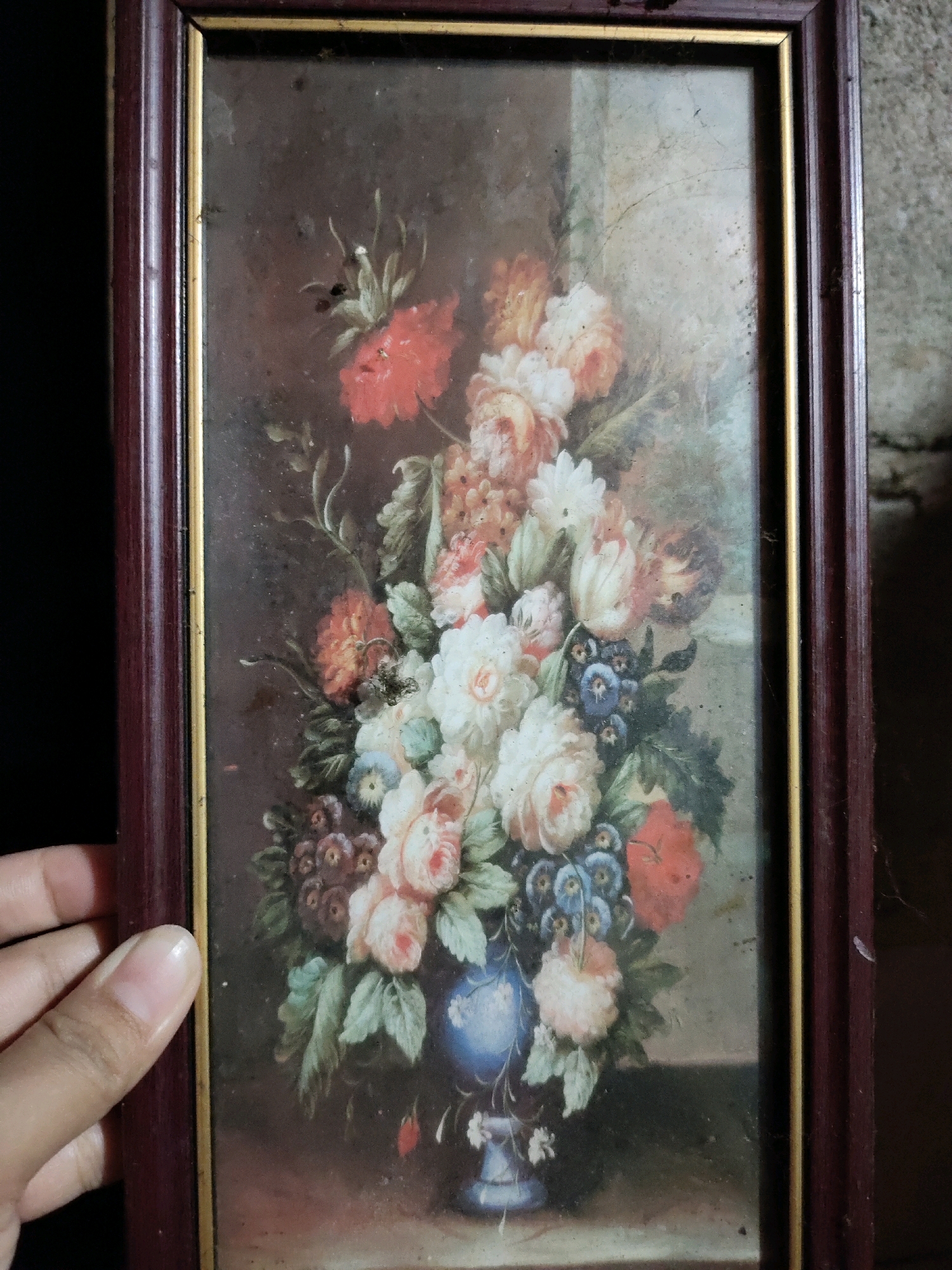 The image shows a framed painting of a floral arrangement. The painting depicts a bouquet of various flowers, including roses, and smaller blue flowers, in a blue vase. The flowers are predominantly in shades of pink, white, and red, with green foliage. The background is dark and muted, creating a contrast with the vibrant flowers. The painting is enclosed in a dark wooden frame with a gold inner trim. The glass covering the painting appears dusty and smudged, with visible scratches and minor damage, suggesting age and wear. The bottom left corner of the image shows a portion of a person's hand, with fingers resting on the frame, indicating someone is holding or displaying the painting. The overall aesthetic is that of an antique still life artwork. The provided context of Matalam, Philippines, does not directly relate to the content of the image itself, as it is a still life painting and not a depiction of a specific location or event. Therefore, details about the city of Matalam cannot be derived from the visual information in the image.