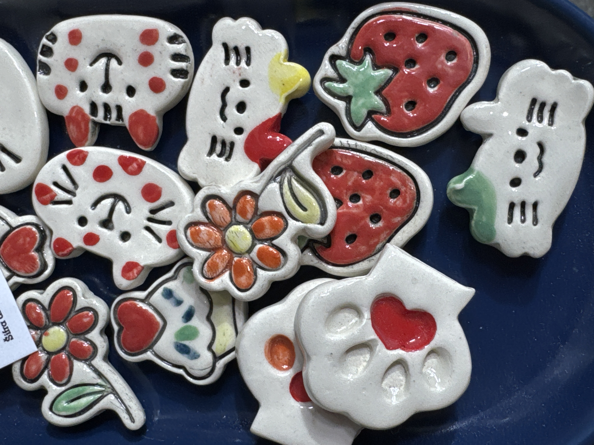 A close-up shows a collection of ceramic decorative elements arranged on a blue surface. The pieces include whimsical animal shapes, such as chicken-like figures and those resembling cartoon animals, along with strawberries and flowers. The items are glazed and painted with vibrant colors like red, orange, yellow, and green, set against a predominantly white background. A small tag with text is visible near the lower left corner, partly obscured. The setting appears to be indoors. The location cannot be determined as Novi Sad, Serbia from this image alone.
