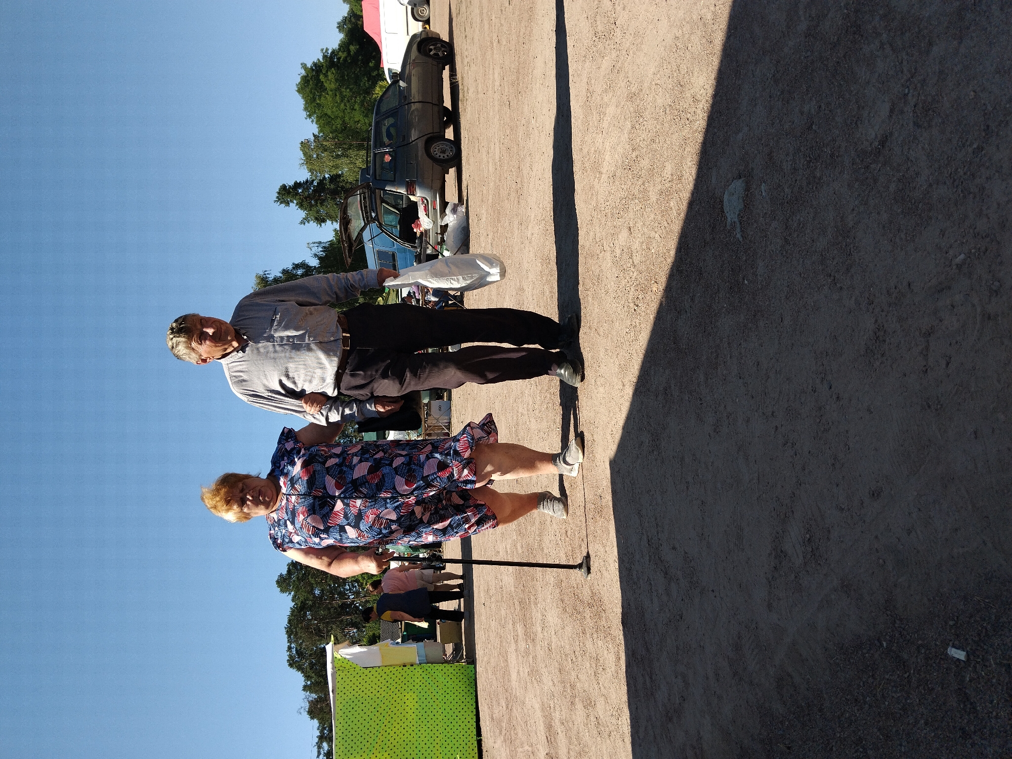 An older woman with blonde hair and a colorful patterned dress is walking with a cane and an older man in a gray collared shirt and dark pants. They are walking on a dirt path with a sparse scattering of gravel. Behind them, there are several stalls with green and yellow coverings, and further back, a dark-colored station wagon with its trunk open, parked next to a white van. The sky is clear and blue, and trees line the horizon. The scene appears to be an outdoor market. The image was taken on June 9, 2024, in Irshansk, Ukraine.