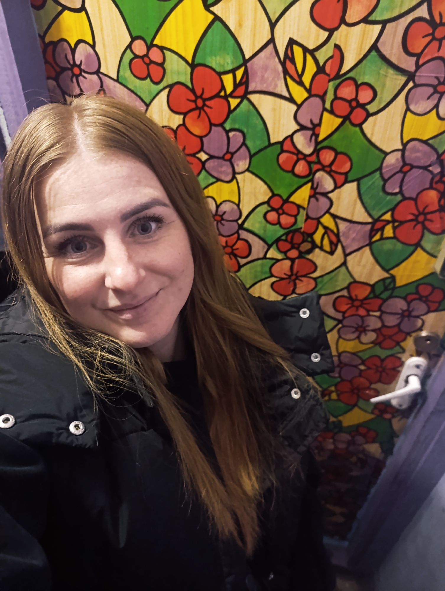 A selfie captures a woman with light brown hair, dressed in a black puffy jacket, indoors. She is positioned in front of a colorful, stained-glass-like pattern featuring stylized flowers in shades of red, purple, yellow, and green, separated by black lines. To the right, a white door handle is visible against the background. A pale blue curtain is draped on the left side of the frame. The location in the city is Unknown, Unknown.