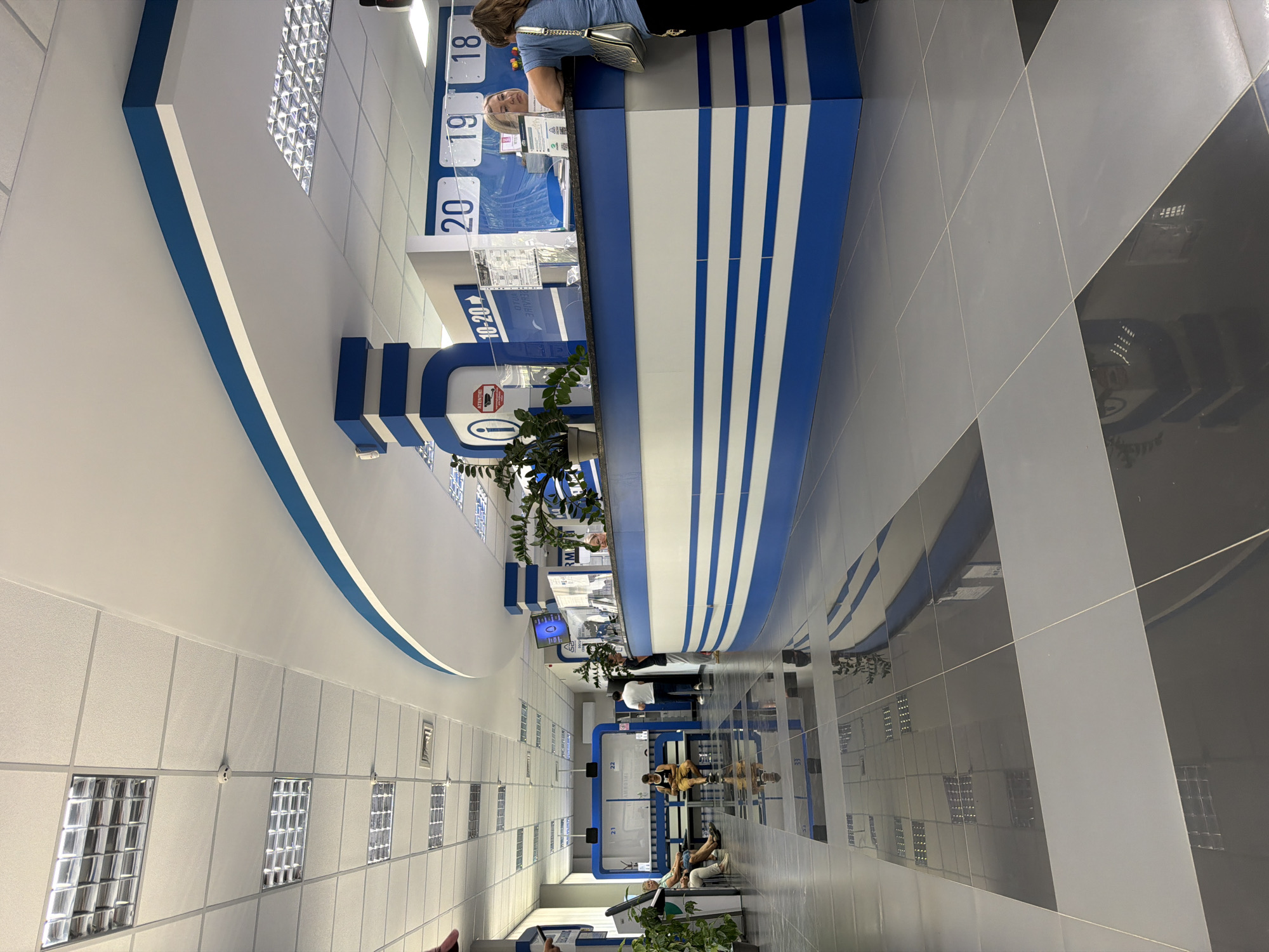 The image depicts the interior of a modern, well-lit office space, likely a public service or administrative building, in Strășeni, Moldova. The environment features a high, white ceiling with a grid of fluorescent lights and ceiling tiles. A curved, blue-and-white partition runs along the right side of the image, separating the service area from the waiting area.
In the waiting area, several individuals are seated on benches. Two women are visible, one in a white top and another in a blue top. A man in a dark shirt and shorts is also seated. The floor is tiled with large, reflective squares in shades of gray and black, creating reflections of the ceiling lights and surrounding architecture.
At the service counter, a woman with blonde hair, wearing glasses, is visible behind a clear plastic barrier. She appears to be interacting with a customer. A woman with brown hair, wearing a blue shirt and a silver handbag, is leaning on the counter, facing away from the camera. Above the counter, digital displays show numbers "20," "19," and "18," likely indicating service queues or office numbers. Plants in potted containers are strategically placed in the area, adding a touch of greenery.
The overall impression is one of an organized and functional administrative facility. The lighting is bright and even, and the clean lines of the architecture suggest a contemporary design.
