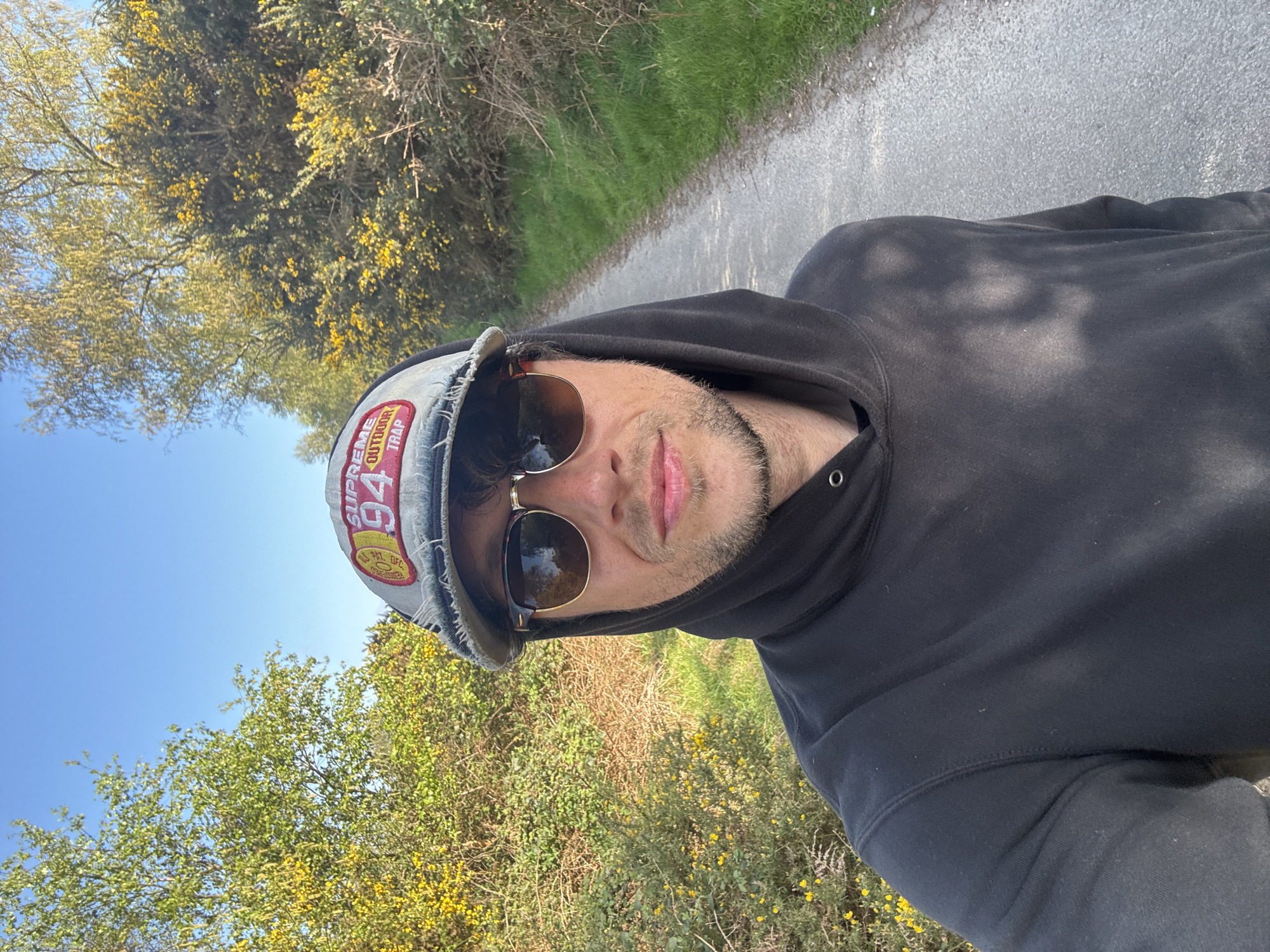 A man wearing a black hooded sweatshirt, sunglasses, and a baseball cap is taking a selfie. He appears to be on a path surrounded by greenery, possibly in East Suffolk, United Kingdom. Yellow flowers are visible on the bushes. The sky is clear and blue. The path is paved.