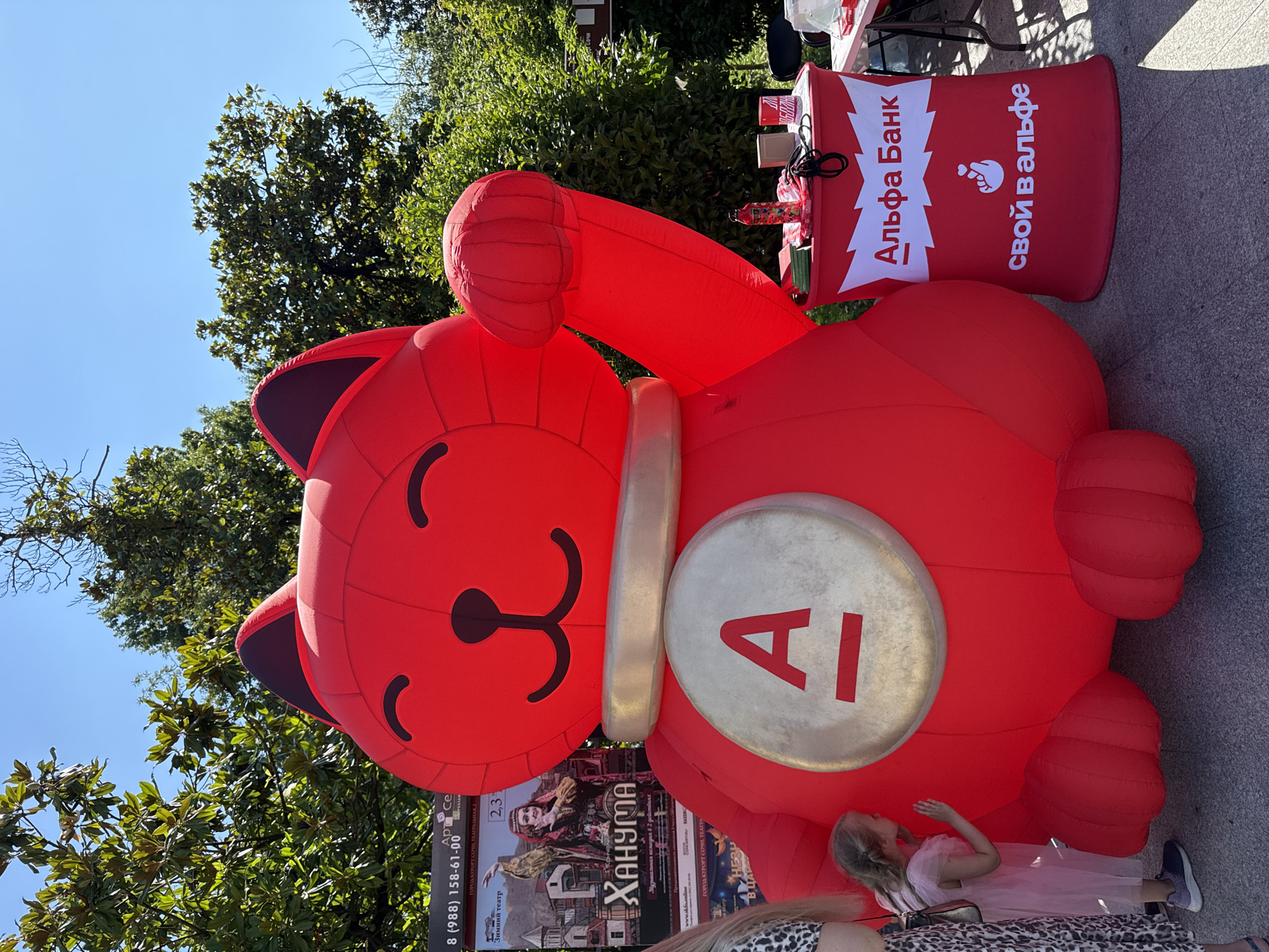 An inflatable red cat statue, resembling a Maneki-neko, stands tall outdoors. A young girl in a pink dress is leaning against the statue, touching it with her hand. To the right of the statue is a red branded table with a logo that says "Альфа Банк" in white text. The table has items on it, including a water bottle, what appears to be a book, and a red cup. In the background, there are trees and a clear blue sky, suggesting a sunny day. There are also posters behind the inflatable cat, one of which advertises "Ханума" a performance with a woman in traditional clothing. This scene appears to be at an outdoor event or festival, possibly in the city of Sochi, Russia, given the Cyrillic text visible.