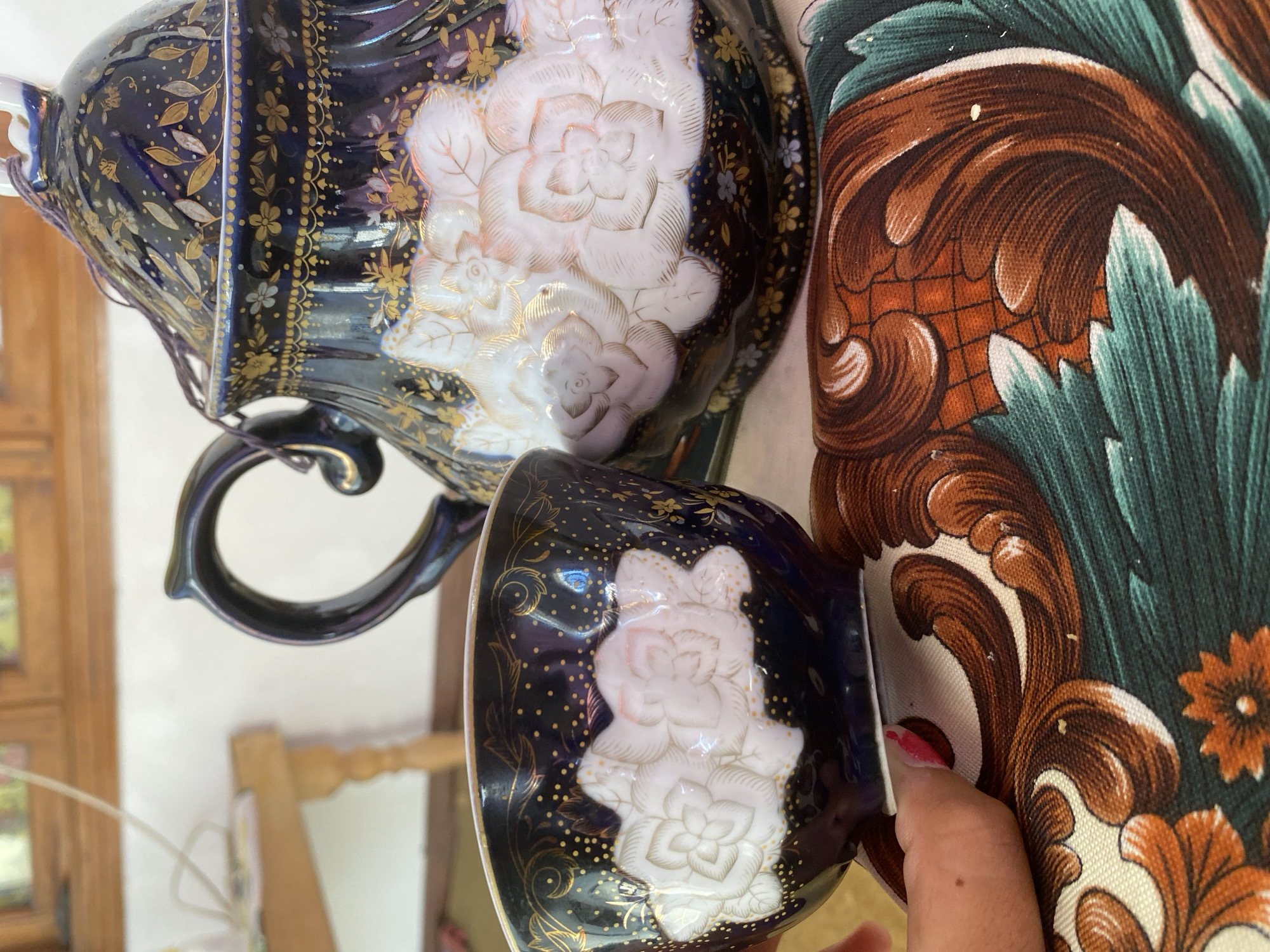 A close-up, eye-level shot focuses on a dark blue porcelain teapot with a lid and a matching cup, both adorned with ornate gold floral patterns and raised white floral designs. The teapot has a dark handle and a decorative lid with a small string attachment. A hand with pink nail polish is visible holding the cup, its fingertip touching the base. The objects rest on a patterned fabric with swirling designs in brown, orange, and teal. In the background, a wooden table with legs is partially visible, along with a window with a wooden frame. The scene appears to be indoors.