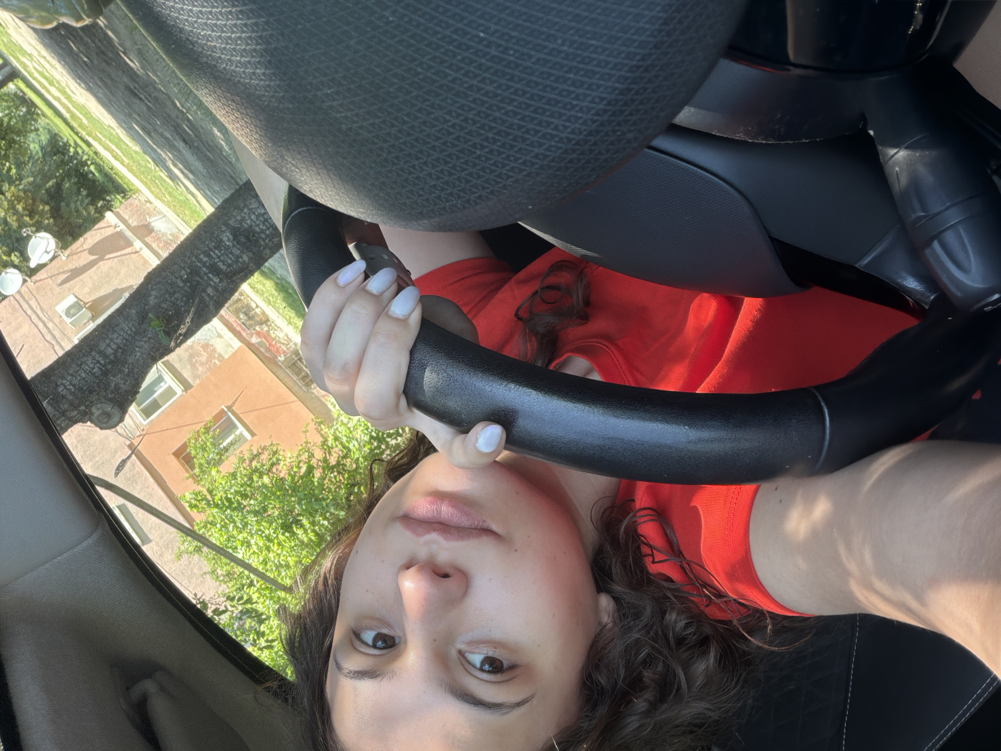 A young woman with curly brown hair and fair skin is in the driver's seat of a car, looking towards the camera. She is wearing a red t-shirt. Her left hand rests on the steering wheel. The interior of the car is dark, with the steering wheel being the most prominent object. Outside the car, a building with orange walls and white windows is visible, along with a tree and greenery. The sun is shining, casting shadows.