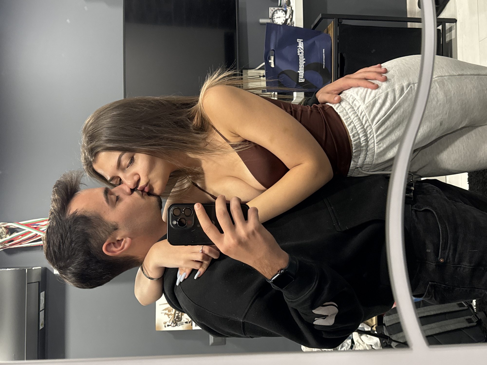 A couple is seen in a mirror selfie, kissing. The man is wearing a black hoodie and black jeans. The woman is wearing a brown top and grey sweatpants. The background includes a television, a blue bag with writing, and shelves. The location is impossible to determine with provided information.