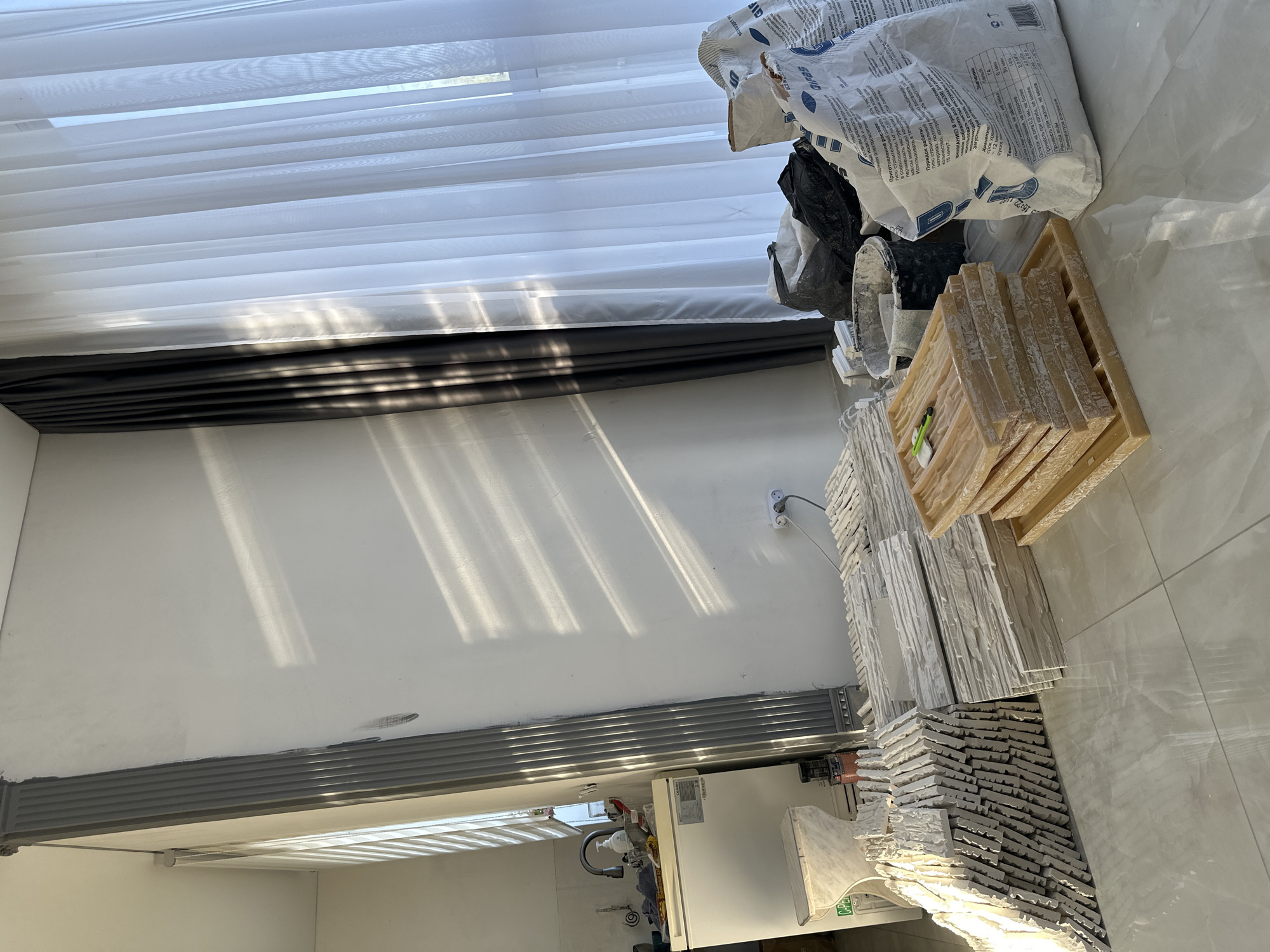 The image shows an interior setting with construction materials. There are bags of building materials stacked on the upper right side. To the side and below them are containers holding tiles. In the lower left corner appears to be the edge of a kitchen counter or cupboard area. The background features a wall with sunlight shining across it. There is no information on the location or people present in the image.