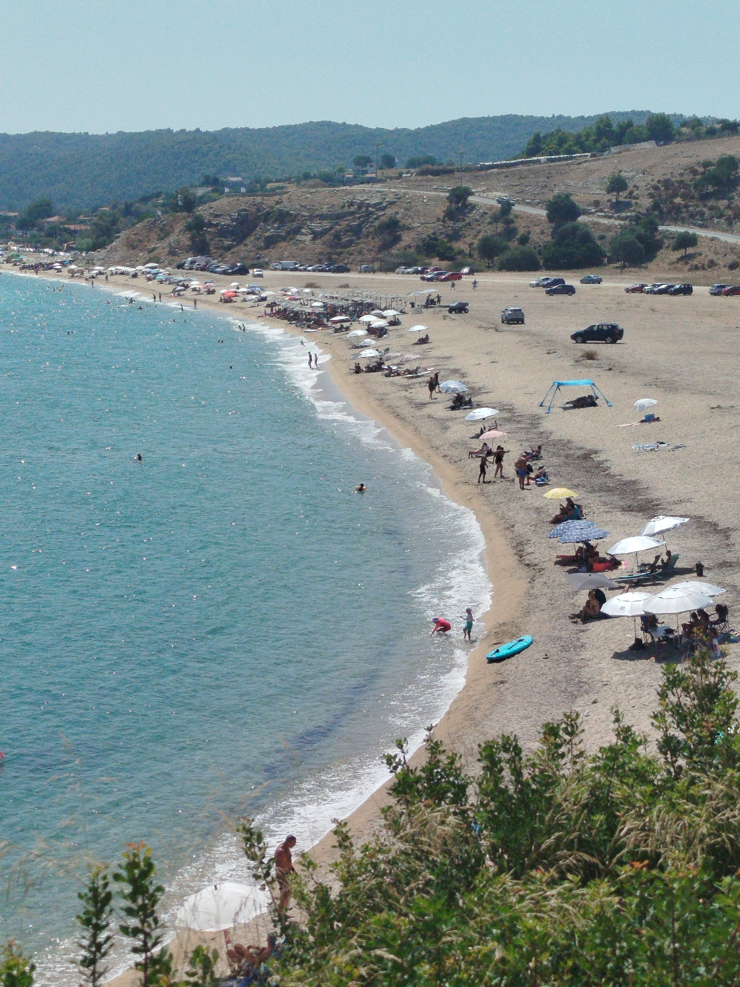 This is an elevated view of a crowded beach on the coast of Sithonia Municipal Unit, Greece. The image captures a section of a sandy beach adjacent to the bright blue Aegean Sea. Numerous beach umbrellas of various colors dot the sand, providing shade for sunbathers and groups of people. Several individuals are visible on the beach, some walking, others relaxing under the umbrellas, and a few are in the shallow water. A small number of people are swimming in the sea, with one person appearing to be wading near the shore. Along the upper edge of the beach, a sandy area is filled with parked cars and a few small structures, suggesting an access point or parking lot. In the background, rolling hills covered with vegetation rise up, with some buildings visible on the slopes. The sea has gentle waves lapping at the shore. The overall scene depicts a vibrant and popular beach destination on a sunny day.
