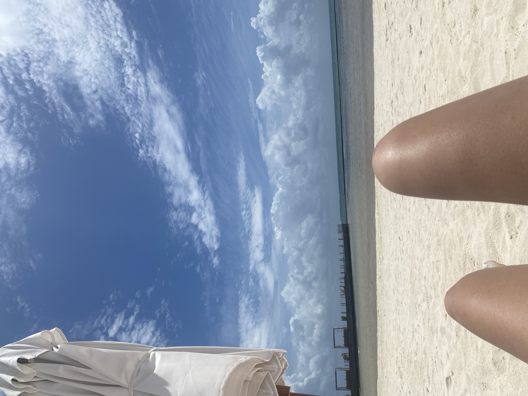 The image depicts a daytime scene at a beach in Marina Maroma, Mexico. The sky, occupying a large portion of the view, is predominantly blue with varying cloud formations. Some clouds are light and whispy, while others are larger and more cumulus-like. The beach itself is sandy and stretches towards the horizon. Parallel to the waterline are dark, evenly spaced structures. To the lower left, a white beach umbrella is partially visible. In the lower right are a person's thighs resting on the sand. One toe is visible.