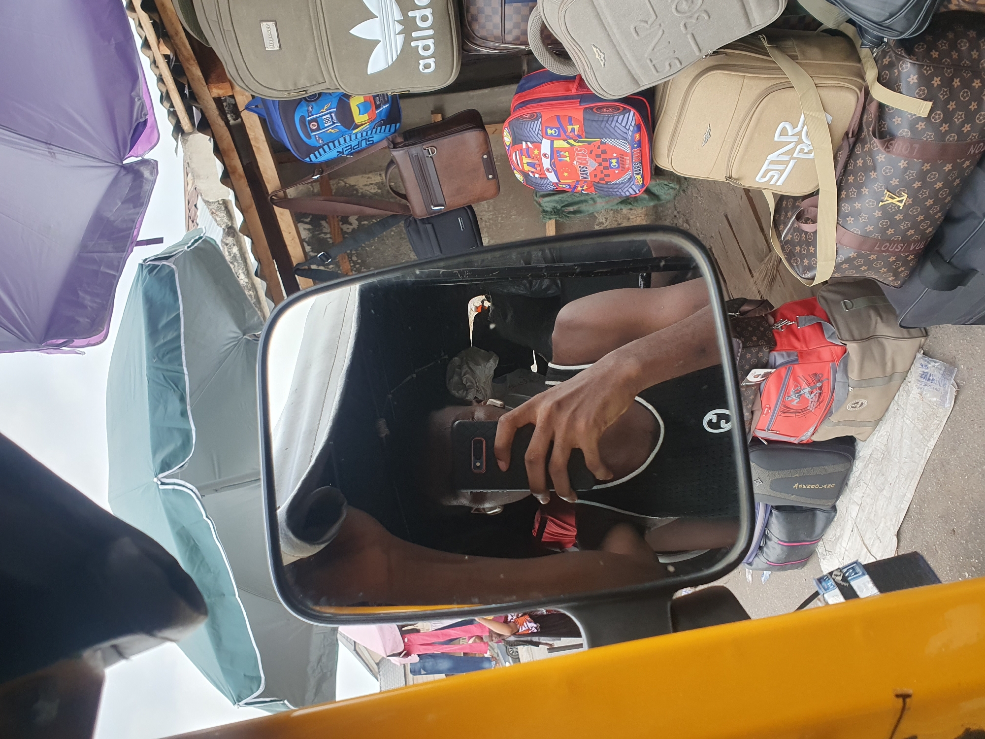 The image presents a vibrant outdoor market scene in Lokoja, Nigeria, observed from the perspective of someone inside or next to a yellow auto-rickshaw (Keke Napep). The central feature is the vehicle's rearview mirror, reflecting a person holding a smartphone to capture the scene or a selfie. The person's arm and torso, clad in a black and white top, are clearly visible in the reflection.
Surrounding the mirror, numerous bags and backpacks are displayed, indicative of a market stall. These include a large beige Adidas bag with its distinctive logo, a blue children's backpack featuring a race car and "SUPER" text, and a red backpack with a similar car theme labeled "KARS KANG." Other bags include a beige duffel bag with "STAR WARS" text, several brown patterned bags resembling Louis Vuitton designs (with visible "LOUSI VUL" text), and a black and grey bag bearing the text "MSENBOLANO." Generic bags in various colors like brown, red, and black are also part of the display.
Above the bags, parts of two large, open umbrellas—one purple and one green—are visible, likely providing shade or shelter from rain. The sky appears bright and overcast, suggesting daytime. Wooden structures and concrete can be seen behind the merchandise. The scene captures the everyday commerce of an outdoor market in a bustling urban environment.