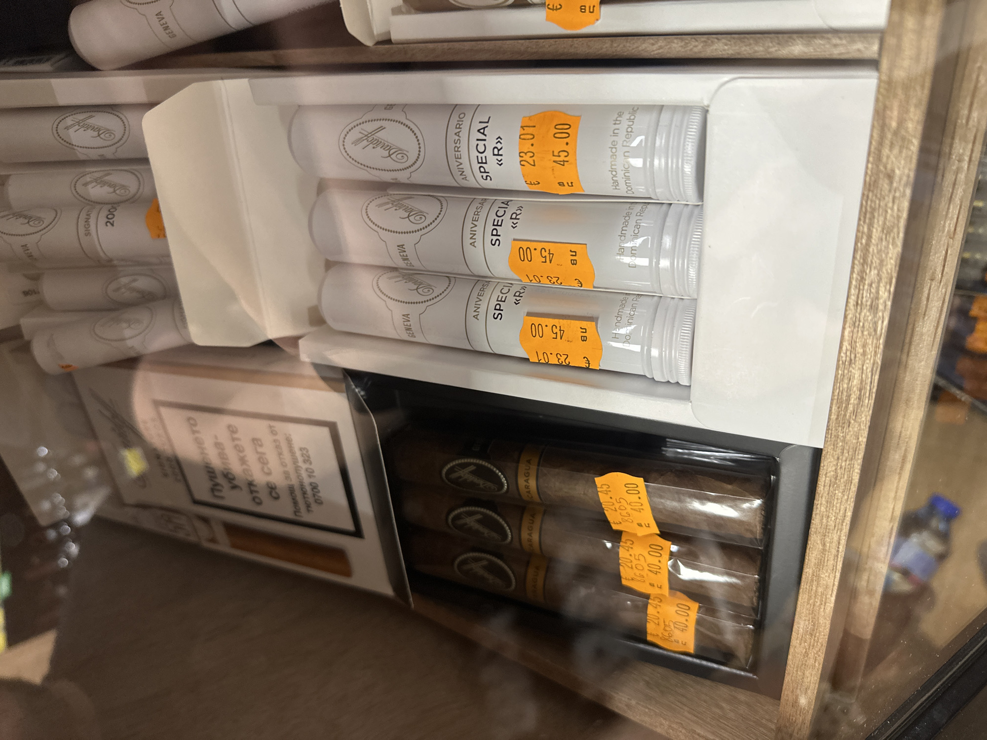 The image shows a close-up view of a retail display case, likely in a tobacco shop or duty-free store. The display features several rows of premium cigars and cigarillos. In the foreground, three individually packaged cigars are visible, each with an orange price tag. The price tags indicate "€ 20.45" and "40.00". Above these, in the middle section of the display, are three white cylindrical tubes, each containing a cigarillo. These are labeled "ANIVERSARIO SPECIAL «R»" and also have orange price tags with "€ 23.01" and "45.00". The tubes are branded with "Davidoff" and "GENEVA". In the background, a pack of cigarettes with Bulgarian health warnings ("Пушенето убива - откажете се сега") is partially visible. The display case has a wooden frame and a glass front, suggesting it's a refrigerated or climate-controlled unit. The overall impression is one of a well-organized, upscale product display for tobacco enthusiasts. The location is identified as Unknown, Unknown.