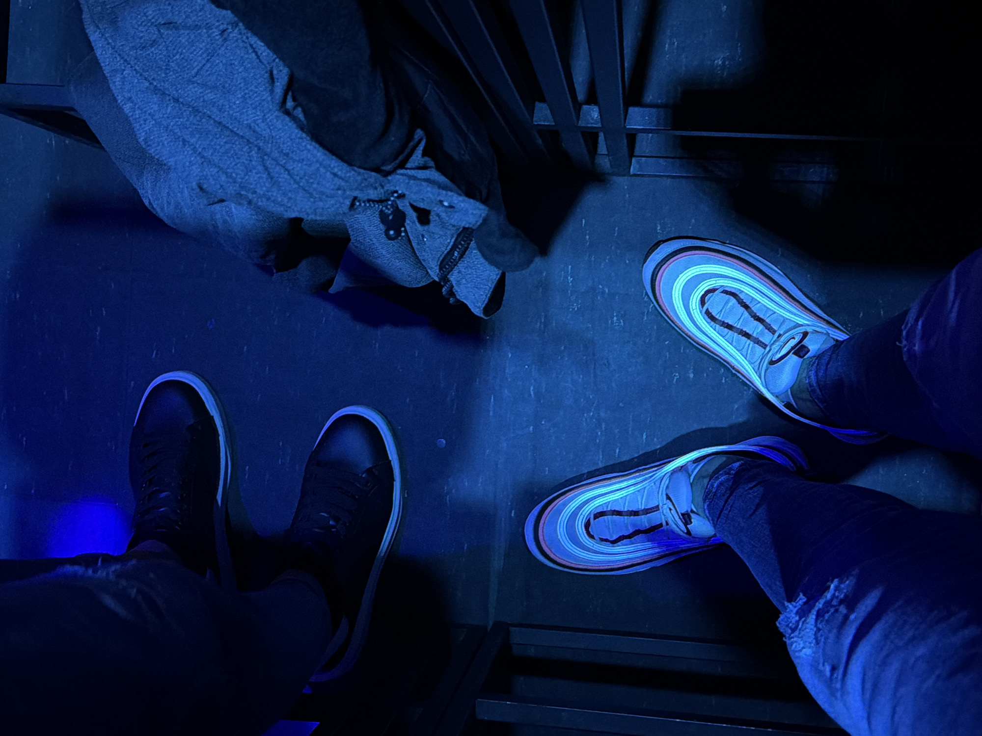 The image presents an overhead view of the lower legs and feet of two individuals in a dimly lit, blue-hued indoor environment.
On the left side, a person wears dark blue jeans and black sneakers with white soles that glow faintly under the dominant blue lighting. A distinct, bright blue light spot is also visible on the floor near their left foot.
On the right side, another person's legs are seen, clad in dark blue jeans, one of which has a ripped knee. They wear light-colored sneakers featuring prominent, multi-colored stripes in shades of pink, light blue, and white. These stripes fluoresce vibrantly, glowing intensely under the blue or ultraviolet (UV) light that bathes the scene.
In the upper left portion of the frame, a folded grey garment or bag, detailed with a zipper and a small dark embellishment, rests on a dark surface, possibly a bench or shelf. Toward the upper right and center, dark, linear structures, appearing to be metal bars or part of a grate, are visible, casting deep shadows.
The floor beneath the subjects is dark and textured, likely concrete or a similar material, reflecting the pervasive blue ambient light. The intense blue illumination and the glowing elements on the sneakers suggest an environment like a club, a themed event, or an art installation designed with UV lighting. No faces, upper bodies, or visible text are present in the image.