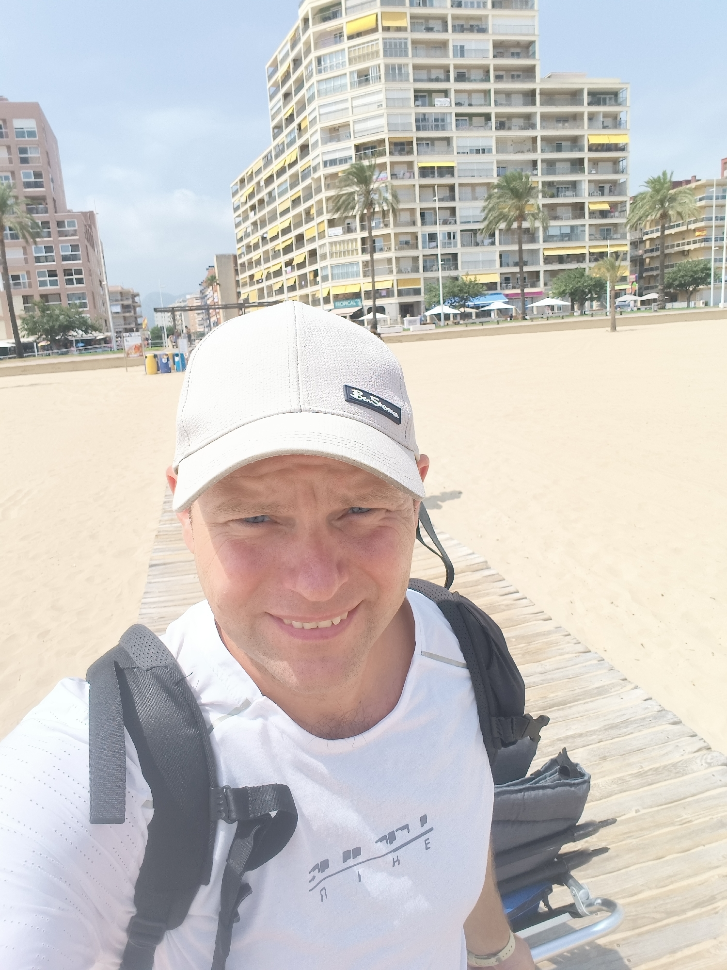 A male subject is positioned in the foreground of the image, facing the camera with a slight smile. He is wearing a white t-shirt, a light-colored baseball cap, and a black backpack. The background reveals a beach scene in Gandia, Spain, with a sandy shore, a wooden boardwalk, and several multi-story buildings. Palm trees are visible between the buildings and on the beach. The sky is clear and bright, suggesting a sunny day. The scene appears to be a tourist area, with beach umbrellas and loungers visible in the distance.