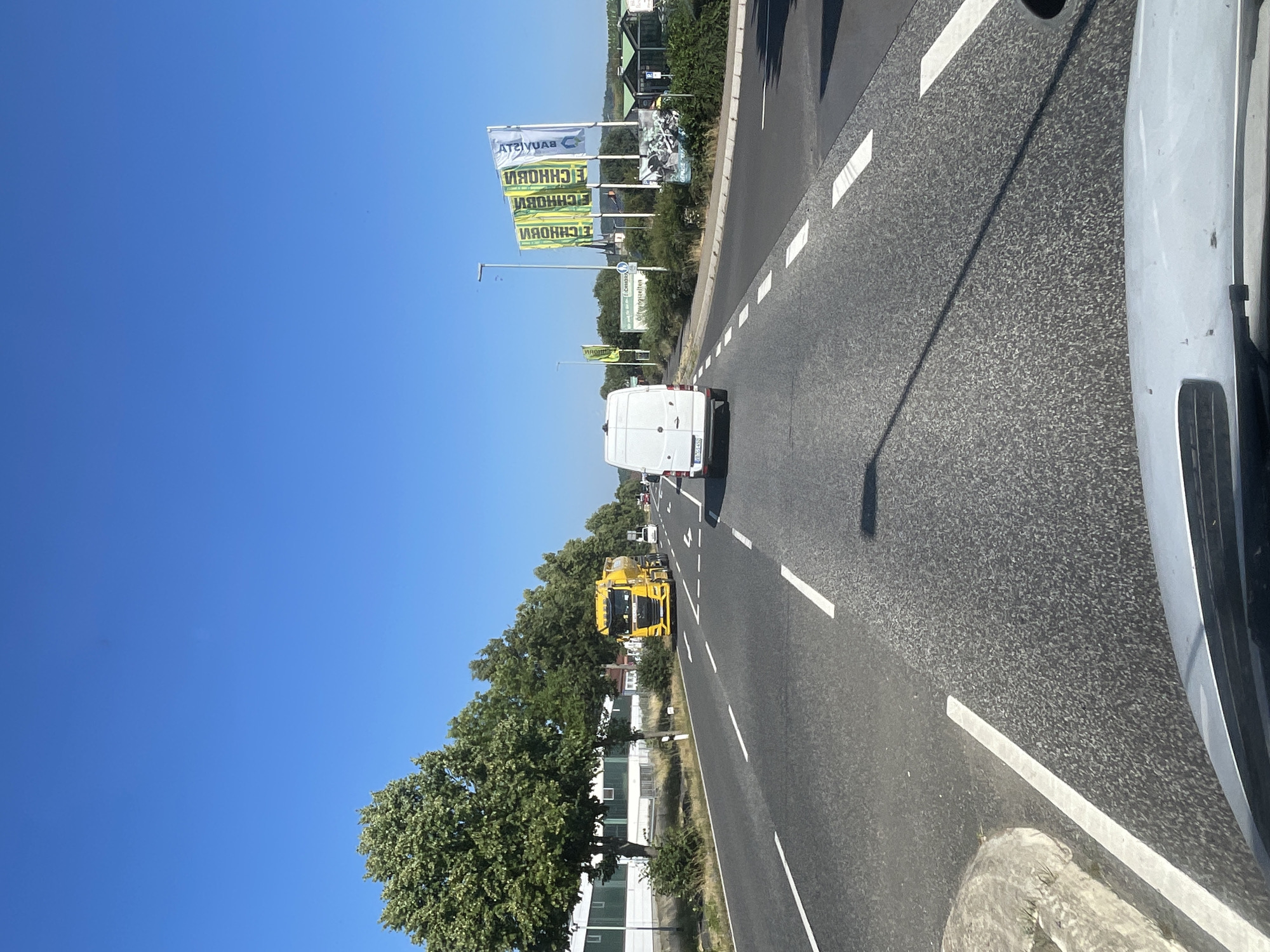The image depicts a sunny daytime scene on a multi-lane road in Wartenberg, Germany. The road has clear white lane markings and asphalt paving. On the left side of the road, there are trees lining the pavement in front of a low-rise industrial building with white walls and a dark roof. Further down the road on the left, a bright yellow tanker truck is visible. In the center of the road, a white panel van is driving away from the viewer. On the right side of the road, several large flags with green and yellow lettering are displayed. The sky is clear and blue. The image appears to be taken from inside a vehicle, looking forward.