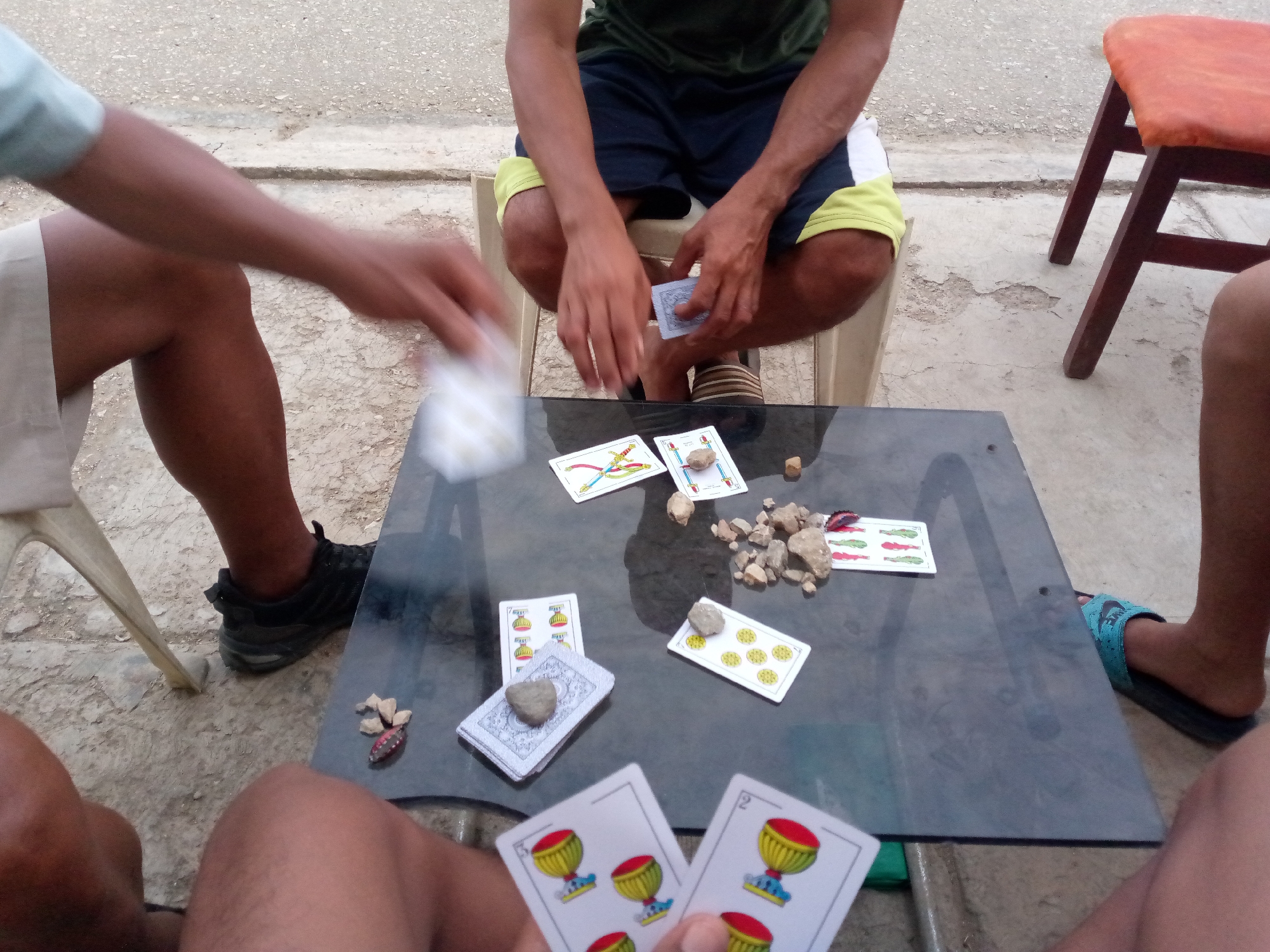 Four individuals are engaged in a card game on a small, dark-colored table. The cards are of a Spanish-suited type. Small stones are scattered on the table. The players are seated on simple chairs. The location is likely outside, given the concrete ground. The city of Juan Griego, Venezuela is known for this game.
