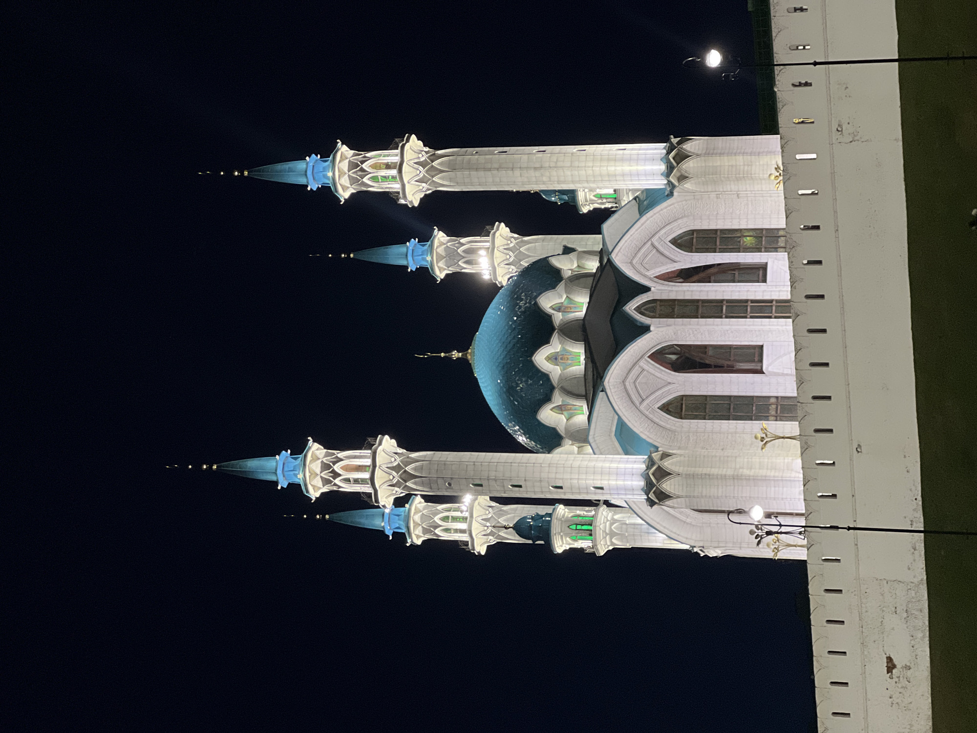 An outdoor night shot of the Qolşärif Mosque in Kazan, Russia. The mosque is brightly illuminated, showcasing its multiple minarets with blue spires and a large blue dome. A portion of a white building or wall with architectural details is visible on the right side of the frame. A lamp post extends into the scene from the building. The background is a dark night sky.