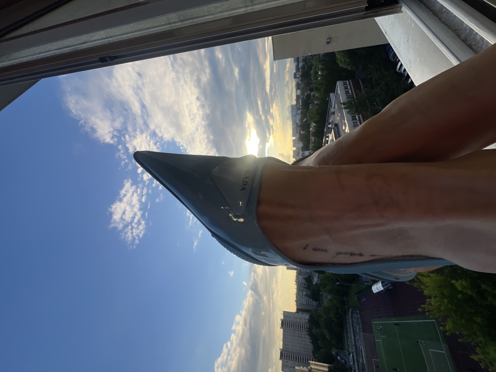 The image is a close-up shot of a person's legs, one adorned with a light blue Prada heel, positioned near an open window overlooking a cityscape at sunset. The setting appears to be Saint Petersburg, Russia, suggested by the architectural style of the buildings. A faint tattoo is visible on the person's ankle. The sky is partly cloudy, with the sun setting behind the clouds, casting a warm glow over the scene. In the background, apartment buildings, trees, and a sports field are visible. The person is leaning against the window frame, with their legs extended. The overall impression is one of relaxation and enjoyment of the evening view.