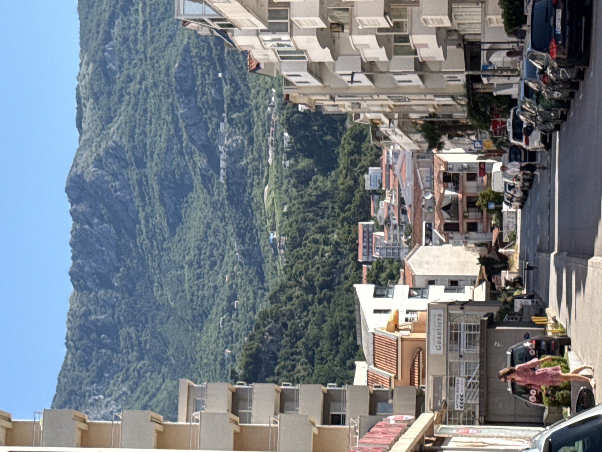 This is a full-frame, eye-level, slightly upward-tilted shot of a street in Budva, Montenegro, on a sunny day with a clear blue sky. The scene features a street lined with multi-story buildings on both sides and a mountainous landscape in the background.
On the left side of the street, a light-colored, multi-story apartment building with balconies dominates the foreground. Its facade is segmented by balconies with white railings, and it has a recessed ground floor with windows and a red railing. Further down the street on the left, a building with orange tiled roofs and a sign reading "Guantiera" is visible. A woman in a pink dress and sunglasses is walking on the sidewalk in front of a dark-colored SUV.
On the right side of the street, a row of white, multi-story buildings with numerous balconies and large windows stretches into the distance. These buildings have a modern architectural style.
The street itself is paved and appears to be a main thoroughfare. Several cars are parked along the right side of the street, and a few vehicles, including a white van and a white SUV, are driving or parked on the street. In the background, a lush green mountain range covered in dense vegetation rises, with some scattered buildings visible on its slopes.
The overall impression is a vibrant Mediterranean street scene, characteristic of coastal towns with a backdrop of natural beauty.