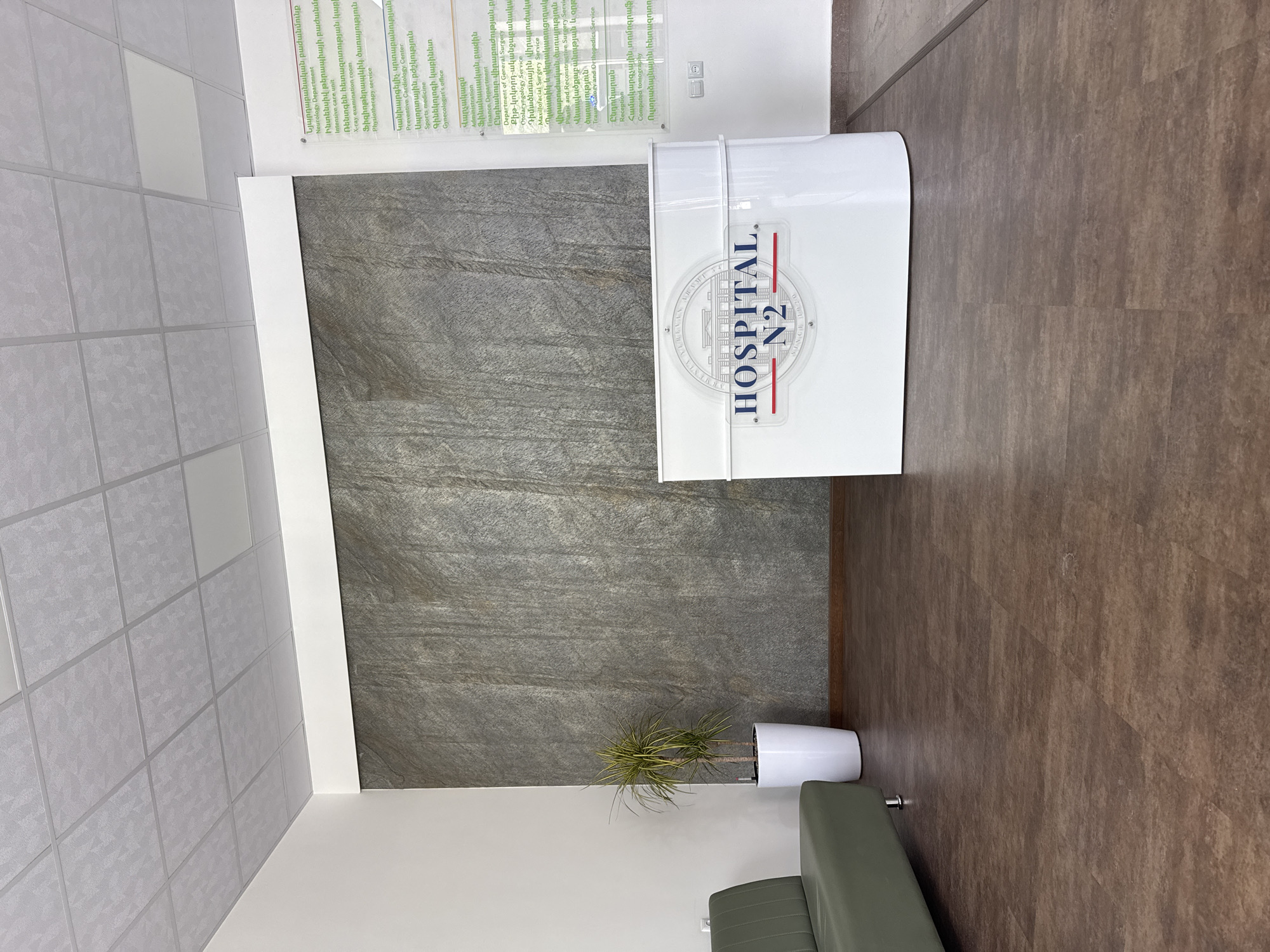 The image depicts the interior of Hospital N2 in Yerevan, Armenia. The scene shows a reception area with a white, curved reception desk displaying the hospital's logo. Behind the desk is a wall with a gray, stone-like texture. To the left, a directory of hospital services is listed in Armenian and English. A potted plant sits near the base of the wall, and a portion of a green seating area is visible at the bottom. The floor is a wood-like texture, and the ceiling has a tiled appearance.