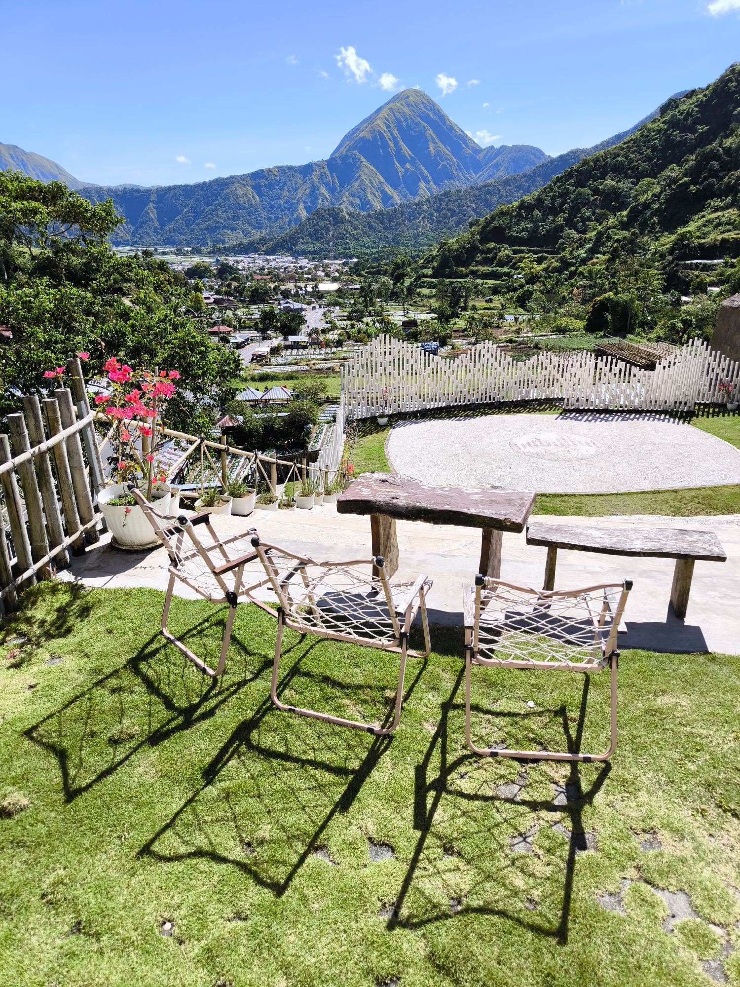 The photograph shows a scenic view in Sembalun Bumbung, Indonesia, featuring a mountain backdrop under a clear, blue sky with scattered clouds. In the foreground, there is a seating area with three tan-colored chairs and a rustic wooden table and bench set on a patch of green grass. A bamboo fence and potted plants with pink flowers border the seating area. Below the seating area, a white picket fence encloses a gravel area with circular patterns etched into the ground. Beyond the fence, a valley stretches out, dotted with buildings and greenery, forming part of the landscape of Sembalun Bumbung. The scene appears to be taken on a bright, sunny day.