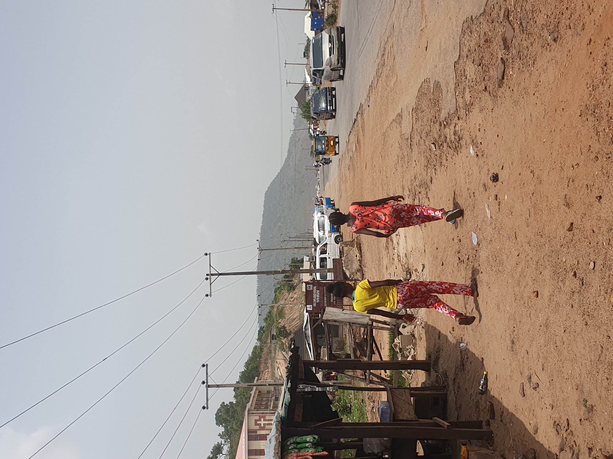 This is a vertically oriented, sunlit street scene in Lokoja, Nigeria, depicting daily life along a main thoroughfare. The paved road, showing signs of wear and potholes, runs from the foreground towards the distant, hazy hills.
In the right foreground, a woman wearing a red floral top and matching pants walks away from the camera on the wide, sandy dirt shoulder. Further left and slightly behind her, another woman in a yellow top and red floral pants stands near a rustic roadside stall, appearing to be engaged in commerce or observation.
The road is active with vehicles, including white vans, darker cars, and yellow and blue tricycles (tuk-tuks) moving in both directions. Utility poles with numerous overhead wires line the left side of the road, extending into the mid-ground. Near the woman in yellow, a wooden stall covered with a dark tarpaulin sells items, possibly foodstuffs in sacks. Above this stall, a brown sign is visible, partially obscured, displaying text including "OGB OGBORIADE FOUNDATION" and "NWA OMIANE," with "SECTION ROADS" also visible.
In the background, a large hill or mountain range is visible under a clear, bright sky, suggesting midday or early afternoon. The overall atmosphere is bright and dusty. The dirt shoulders are strewn with litter, including plastic bottles and papers, indicating a busy area.