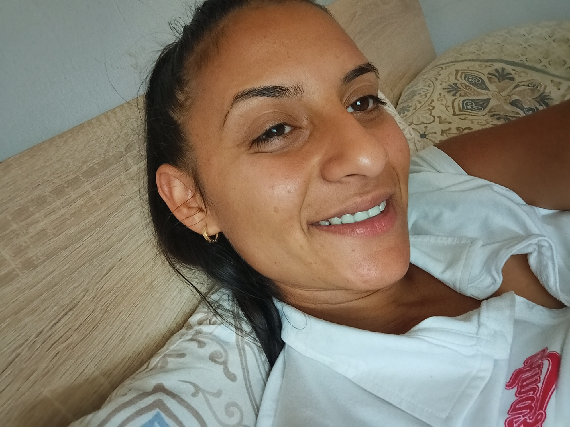A woman is lying down on a bed, looking at the camera. She has dark hair pulled back and is wearing a white collared shirt with a red logo on the chest. She is wearing a small gold hoop earring in her left ear. The background shows a wooden headboard, a light blue wall, and a patterned pillow. The lighting suggests it is daytime. The location is a bedroom in Yambol, Bulgaria.
