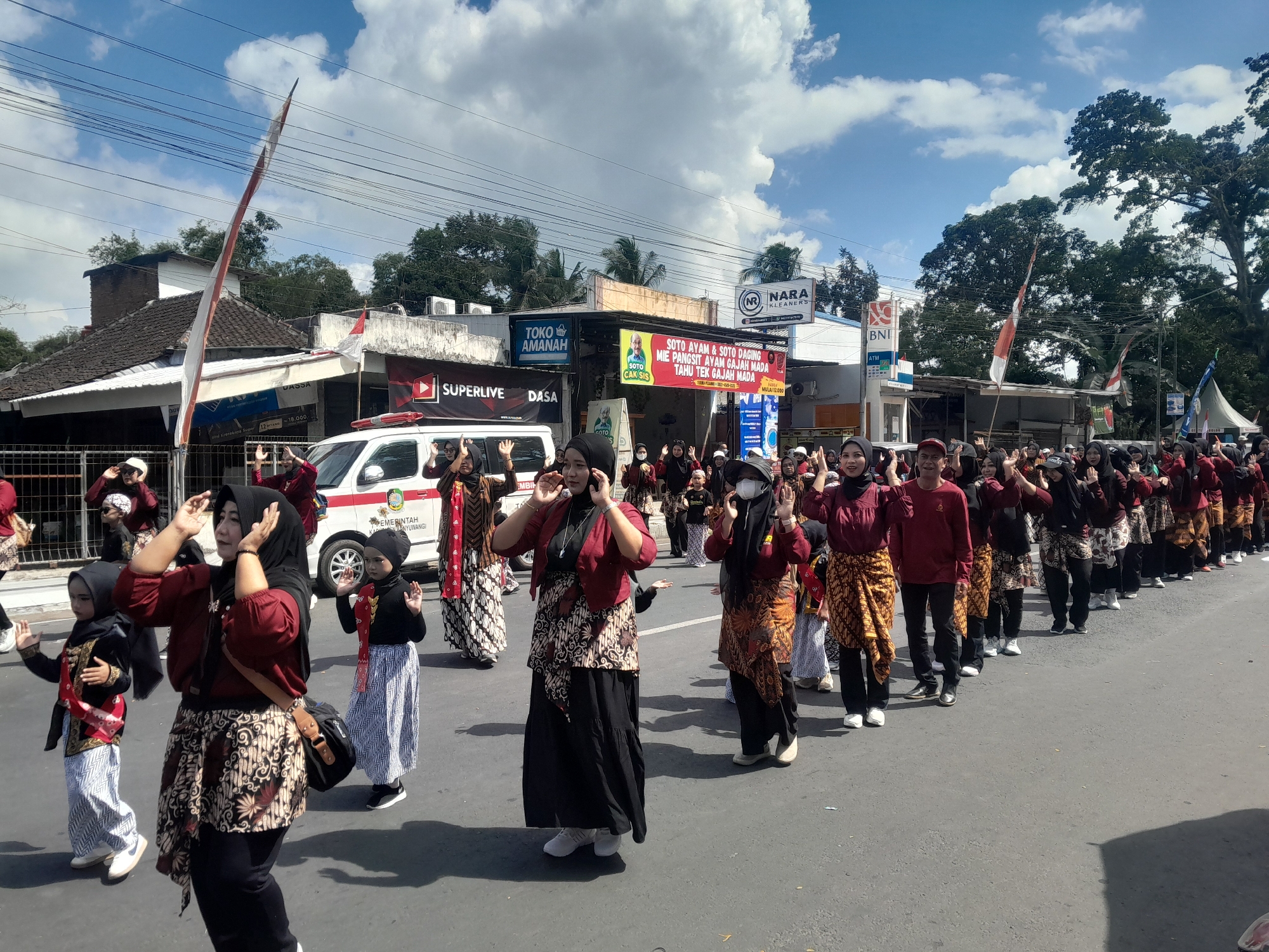 This is a medium shot, eye-level, outdoor photograph taken on a sunny day, showing a procession or parade moving along a paved street. The street appears to be lined with buildings on the left side, including shops and what looks like a government building. The buildings have various signage, some in Indonesian, and a white van is parked on the street. A tall flagpole with a red and white flag is prominently visible on the left, leaning slightly.
The main subjects are a group of people, predominantly women and girls, dressed in traditional Indonesian attire. Many are wearing maroon tops and batik skirts or trousers, with some also wearing headscarves. They appear to be performing a synchronized dance or movement, with many raising their hands to their heads. The children in the front are dressed similarly to the adults, some in black tops with red and white sashes and patterned skirts. The procession extends down the street, with a large number of participants visible.
The background includes lush green trees, a blue sky with fluffy white clouds, and power lines strung across the street. The overall atmosphere suggests a cultural event or celebration taking place in a semi-urban or town setting. The location is identified as Genteng Kulon, Indonesia, indicated by the presence of Indonesian language in the signage and the cultural context of the attire and event.