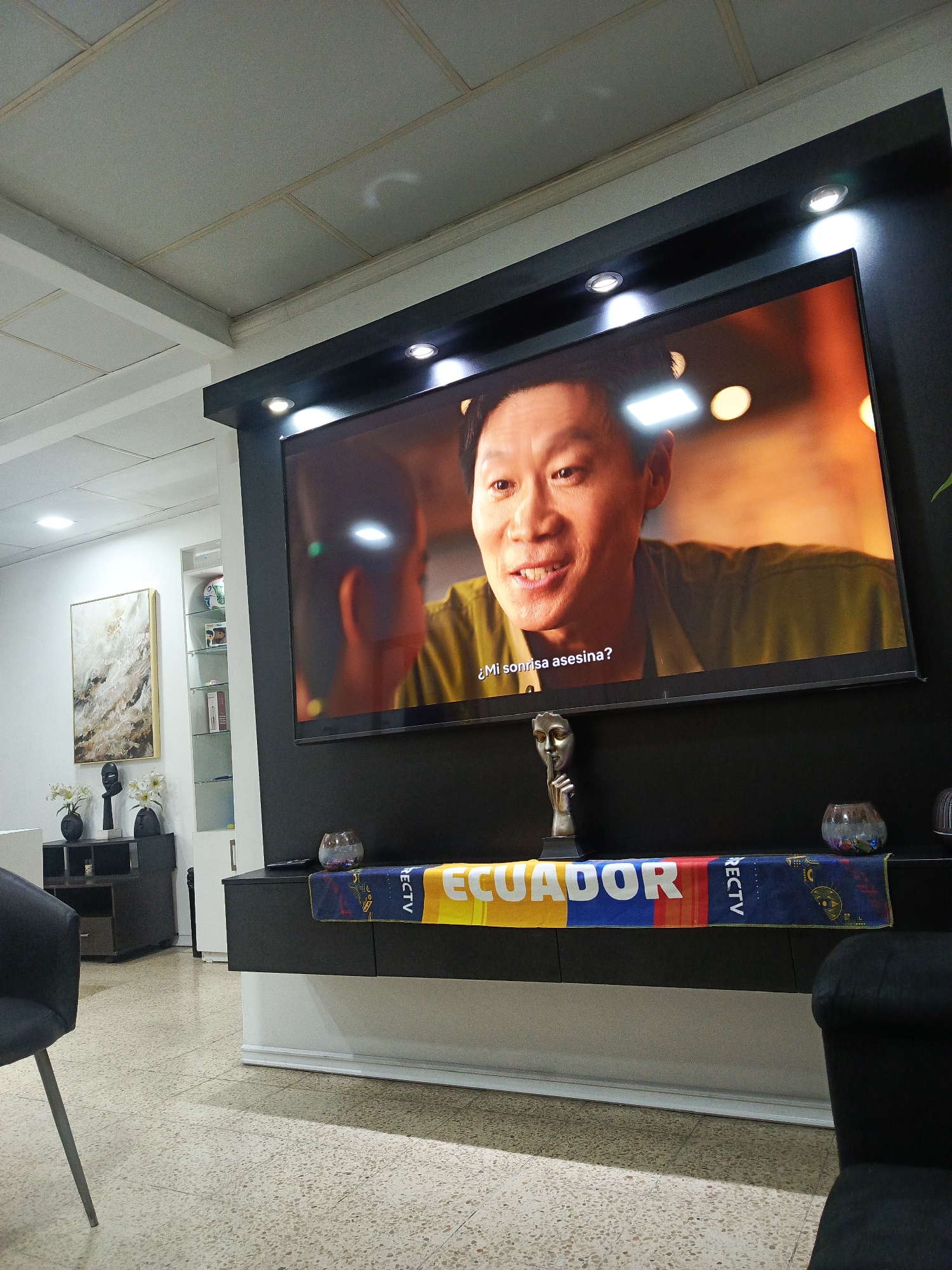 The image depicts an indoor living room area, likely a home or office. A large flat-screen television is mounted on a black wall, displaying a close-up of an Asian man's face with subtitled text below. The television is situated above a long, black media console that supports several decorative items, including a silver bust, a small glass bowl, and a colorful banner that reads "ECUADOR" in large letters, interspersed with the "DIRECTV" logo. The room has a textured off-white ceiling with recessed lighting. To the left of the television, a framed piece of abstract art hangs on a white wall, with a black decorative item and two vases with white flowers below it. Further to the left, a portion of a black armchair is visible. The flooring is a light-colored terrazzo. There are no people clearly visible in the scene, only the man on the television screen. The environment appears tidy and modern. The mention of Guayaquil, Ecuador is not directly supported by visual evidence within the image itself, but the "ECUADOR" banner strongly suggests a connection to the country.