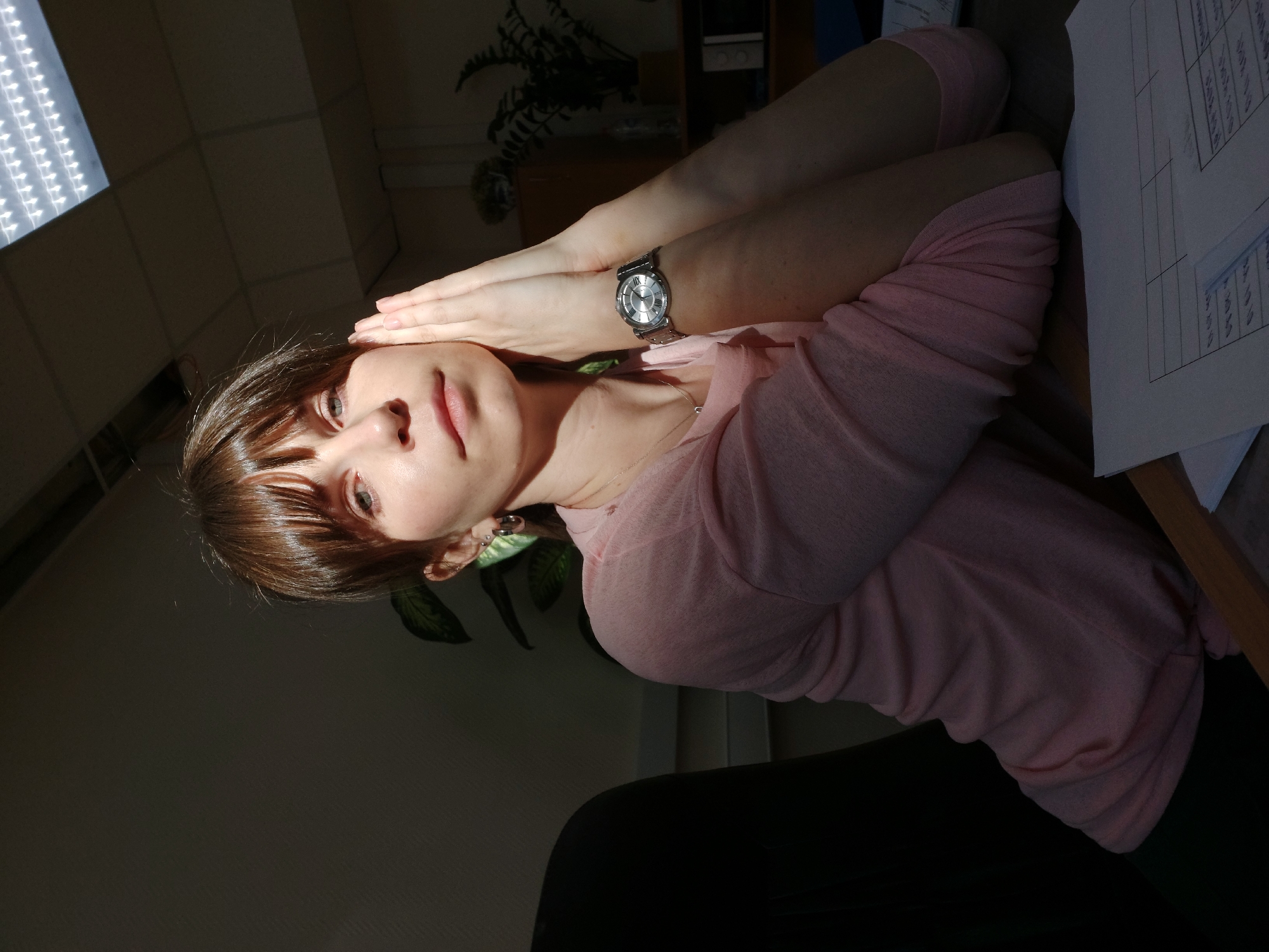 A woman is sitting at a desk. She has brown hair and is wearing a pink shirt and a silver watch. Her hands are raised to her forehead. A sheet of paper is on the desk in front of her. A plant is visible in the background. The location is Chaikovsky, Russia.