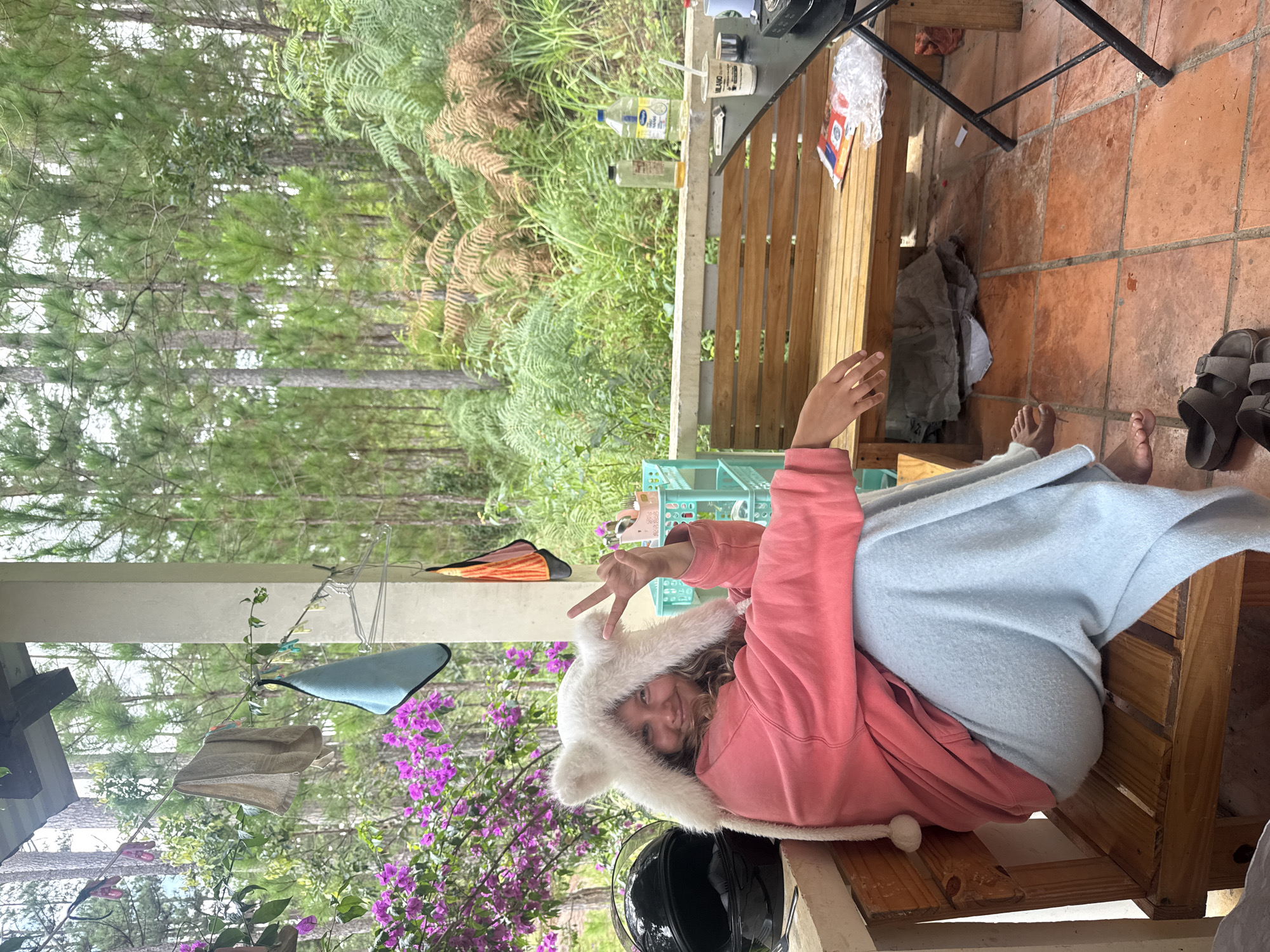 A young woman is sitting on a wooden bench on a porch in Đạ Cháy, Vietnam. She is wearing a fluffy white hat with ear-like protrusions and a pink hoodie. A light blue blanket is draped over her legs. Her right hand is raised, forming a peace sign with two fingers. Behind her, a black motorcycle helmet is visible. The porch has a tiled floor and a wooden railing. In the background, lush green trees and ferns are abundant, characteristic of the Đạ Cháy region. A wooden bench and a black table are also present on the porch. Several bottles and a cup are on the table. A clothesline with towels is hanging nearby.
