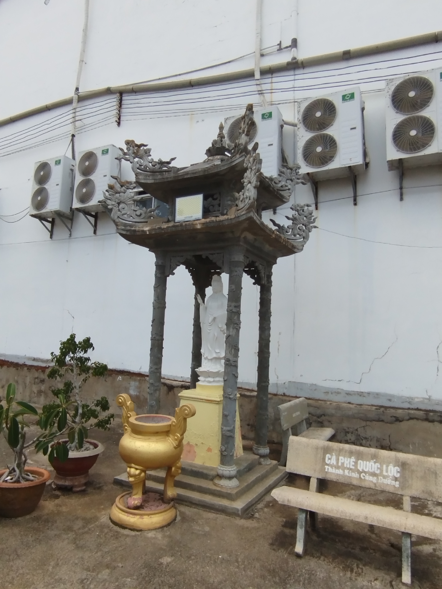 The image captures an outdoor scene in Phú Lộc, Vietnam. A small Buddhist shrine with a white statue of a deity stands prominently. The shrine has a gray, ornate roof and columns. Adjacent to the shrine are a golden incense burner and several potted plants. In the background, a white wall is lined with multiple air conditioning units. A stone bench with Vietnamese script, "CÀ PHÊ QUỐC LỘC THÁNH KINH CÚNG DƯỜNG," sits nearby. The ground is paved with gray concrete.