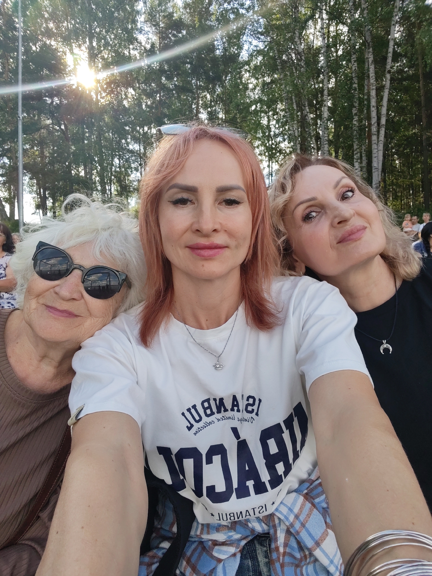 In this media file, three women are captured in a close-up, outdoor selfie. The woman in the center, with pink-tinted hair, is taking the photo. To her left is an older woman with white hair, wearing large, dark, cat-eye sunglasses. To the right of the central woman is another woman with blonde, curly hair. All three appear to be attending an outdoor event.
The background features a line of trees, suggesting a park or wooded area. Sunlight streams through the trees from the upper left, creating a bright, hazy effect with lens flare, indicating it is daytime, likely late afternoon or early evening. The overall mood seems cheerful and relaxed, with the women smiling or looking at the camera.
The central woman wears a white t-shirt with dark blue text that reads "ISTANBUL 21" in a smaller font above larger, bolder text that appears to be "RACKET" or a similar word, though the full word is not entirely clear. She is also wearing a necklace with a crown pendant. The woman on the right wears a black t-shirt and a necklace with a crescent moon pendant. The older woman on the left wears a brown, ribbed top. The lower part of the image shows the central woman's arm and a blue and white plaid shirt or blanket draped over her lap. The presence of other people visible in the background suggests a public gathering.
Based on the provided location context of Chaikovsky, Russia, this scene likely depicts a local event, concert, or festival taking place in the area.