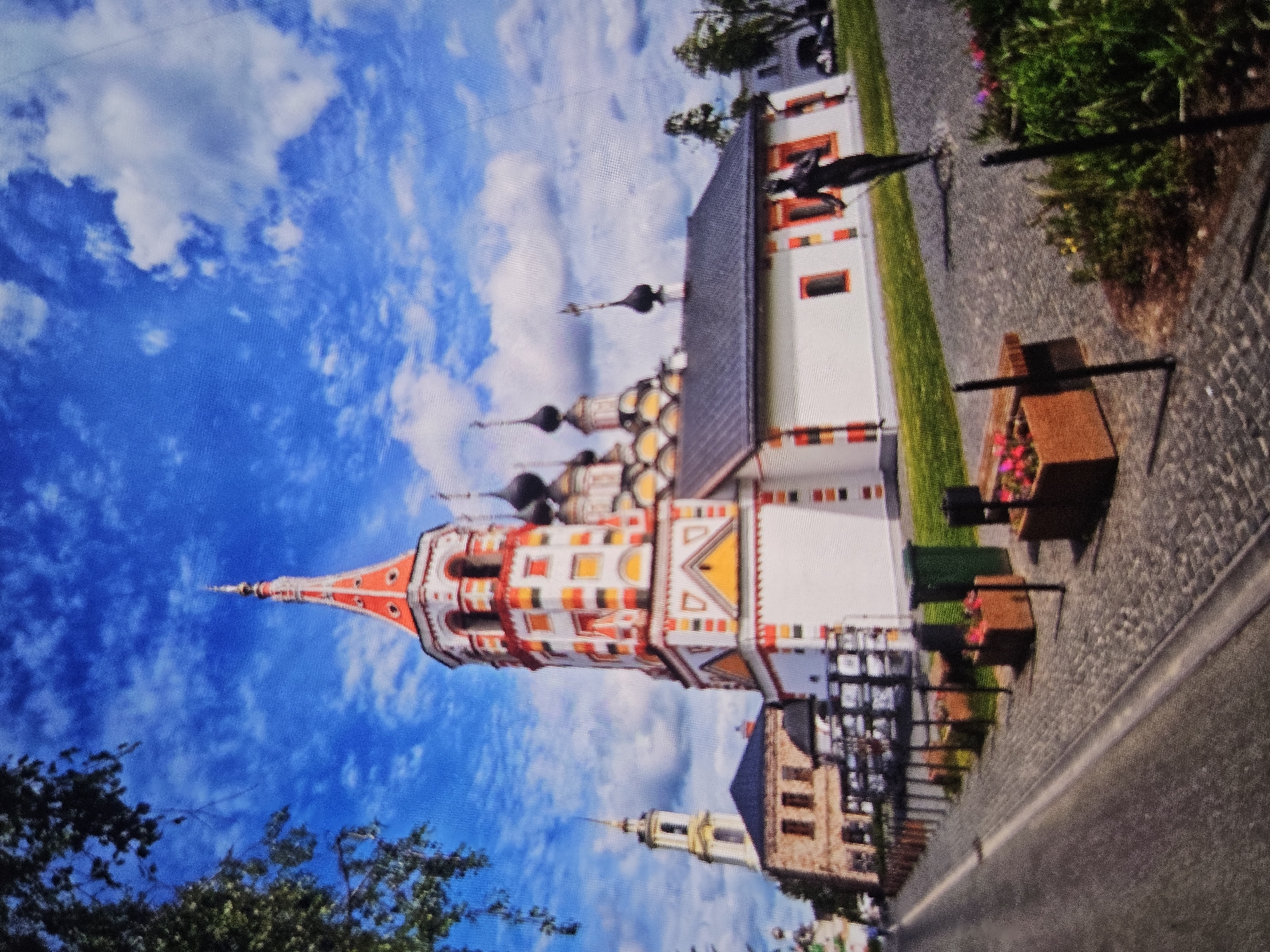 The image shows the Church of the Resurrection in Vladimir, Russia. It is a colorful, ornate church with white walls and a tall, multi-tiered bell tower adorned with red and yellow decorative elements. Several smaller onion domes are visible on the main church building. In the foreground, a paved walkway lined with planters and a bench leads towards the church. A dark statue of a figure holding a sword stands to the right of the walkway. The sky above is a vibrant blue with scattered white clouds. A road is visible to the left, with a tree branch arching into the frame from the top left corner.