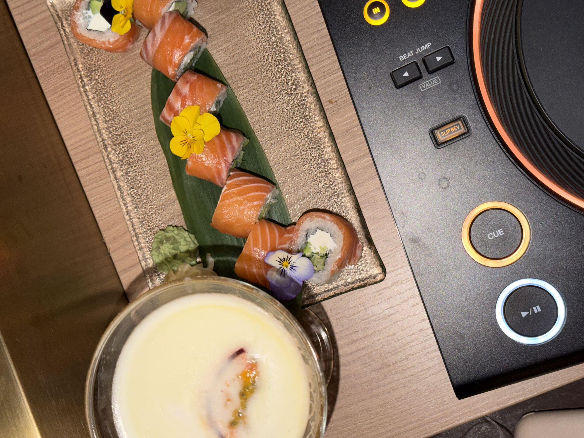 The image shows a wooden table with a plate of sushi and a glass of white liquid, likely a cocktail, placed next to a black DJ controller. The sushi, adorned with small edible flowers, consists of salmon rolls with a creamy filling, possibly cream cheese and avocado. The cocktail has a flower garnish and a light foamy texture. The DJ controller has illuminated buttons labeled "CUE," "PLAY/PAUSE," "BEAT JUMP," and "SLIP REV." The location in Budva, Montenegro is not identifiable from the information in the image.