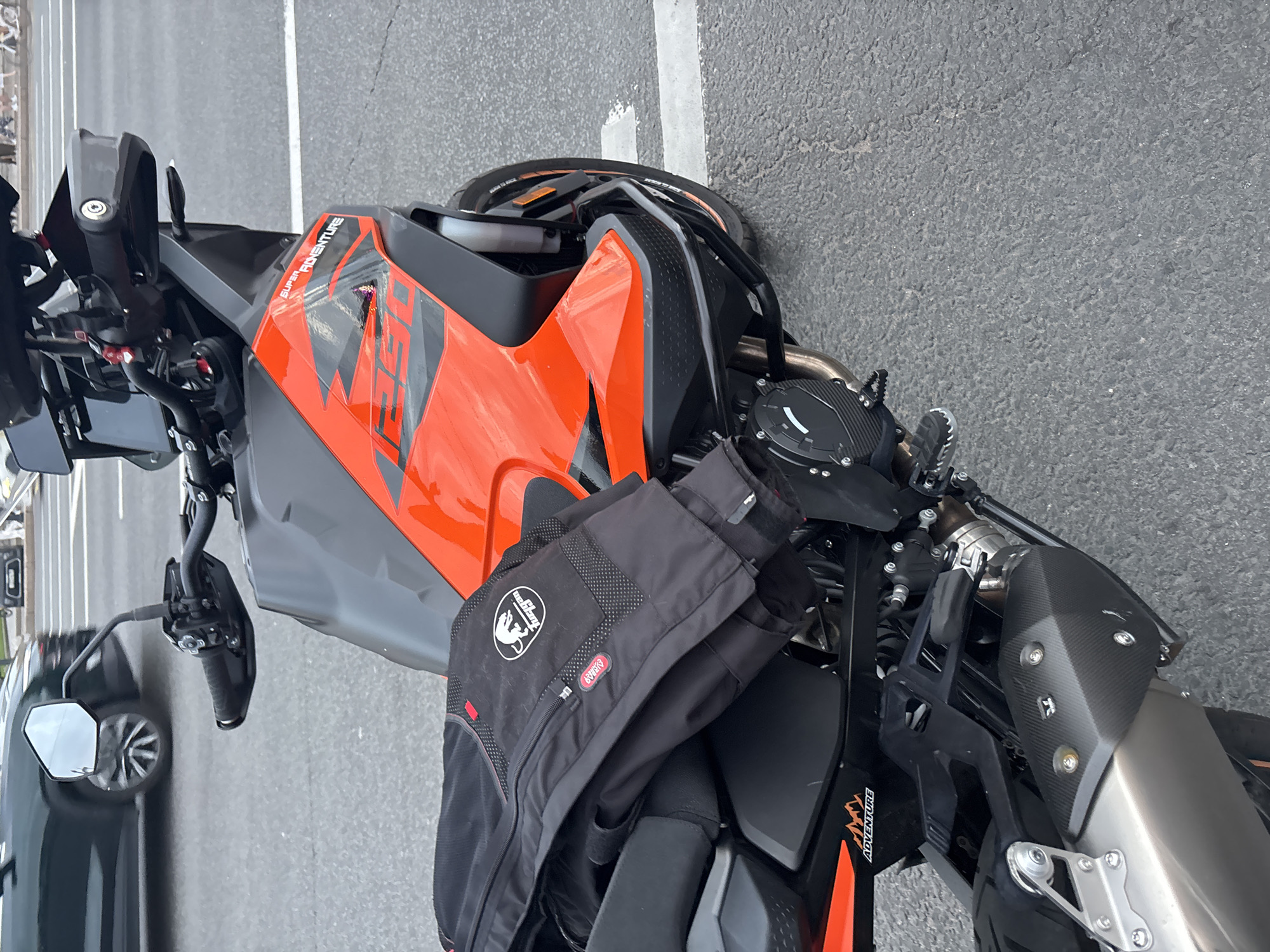 A medium shot captures an orange and black KTM Super Adventure motorcycle parked on an asphalt road in Saint Petersburg, Russia. A black motorcycle jacket is draped over the seat. The motorcycle has a prominent orange fuel tank with black graphics and the text "SUPER ADVENTURE 1290" visible. The front of the motorcycle features a black windscreen and handguards. In the background, a dark green car is partially visible, driving on the road, and a few pedestrians are seen in the distance. The ground is marked with white lines, suggesting a street or crosswalk. The overall scene is a street-level view of a parked motorcycle in an urban environment.