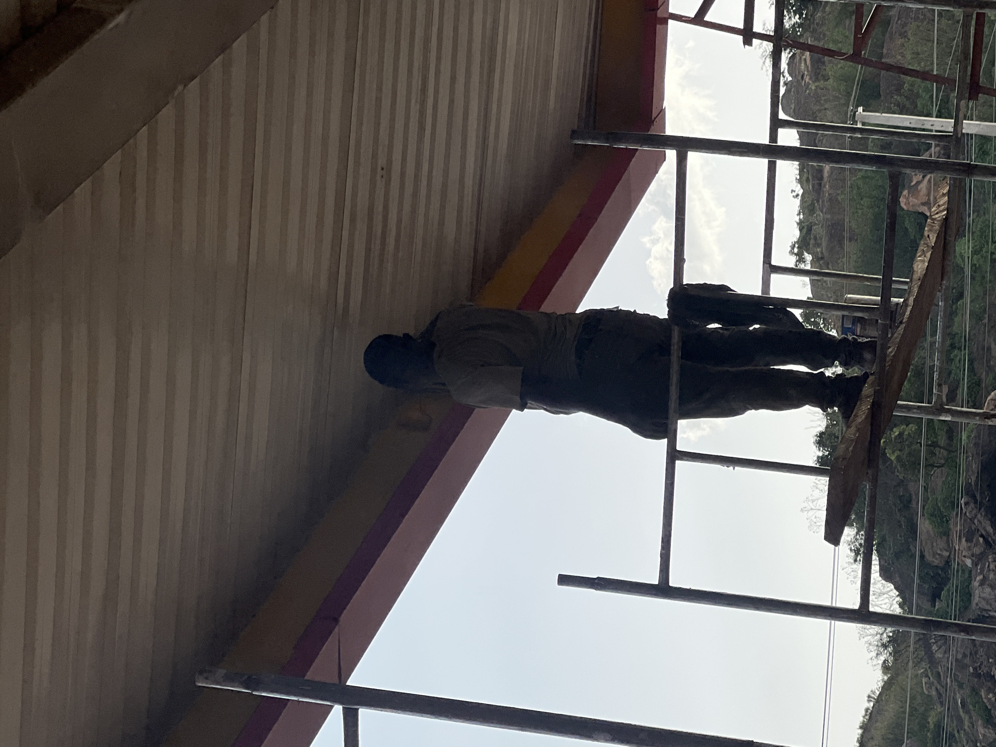 A worker on scaffolding appears to be repairing or maintaining a building's awning in Gura Top, Nigeria. The building's underside has a striped pattern, and the awning is supported by a yellow and red beam. The surrounding environment includes rocky terrain and green vegetation.