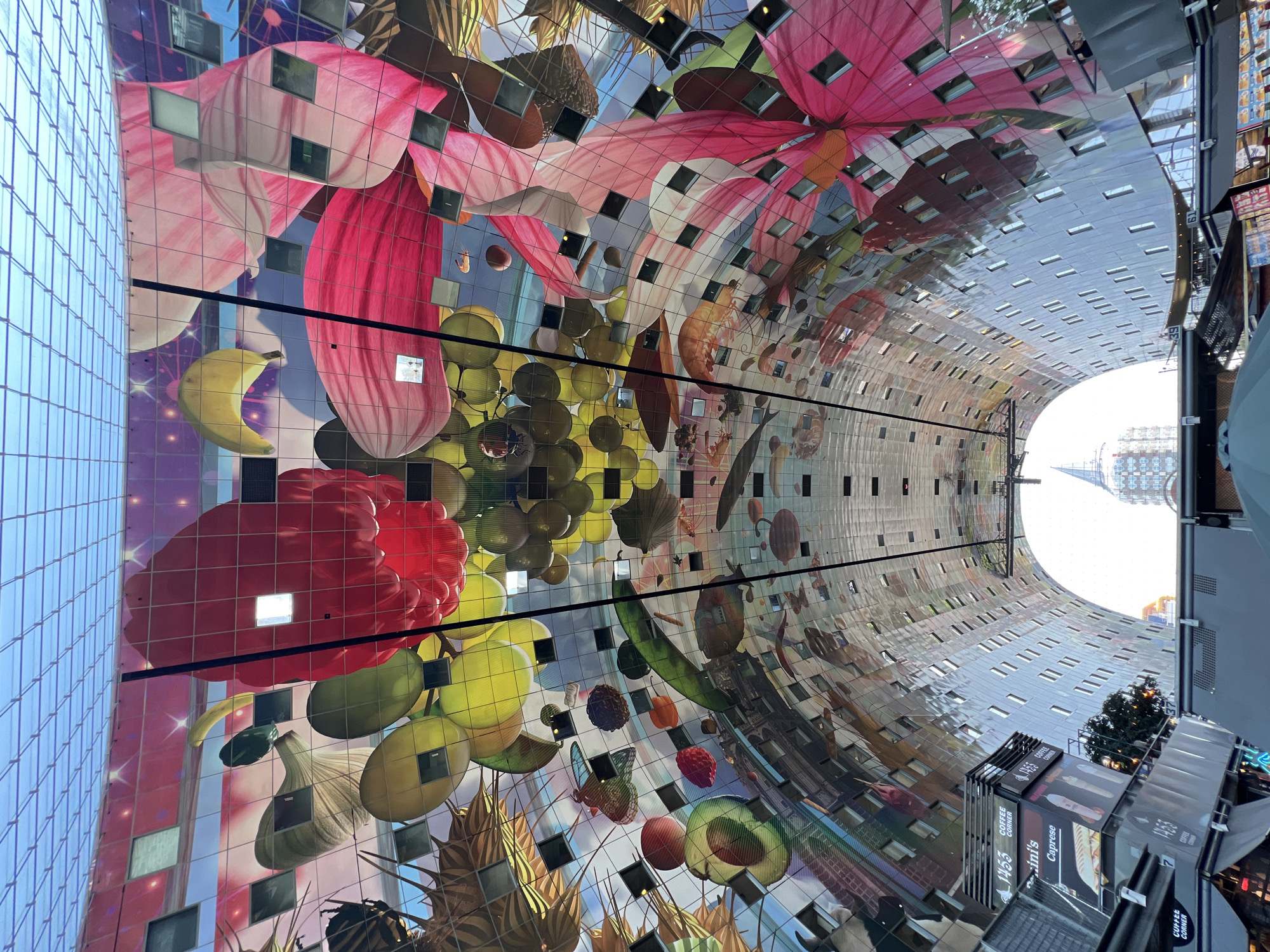 The image shows the interior of the Markthal in Rotterdam, Netherlands, a unique architectural landmark. The ceiling of the Markthal is a massive, curved artwork depicting oversized fruits, vegetables, and insects, creating a vibrant and surreal visual experience. Below the artwork, the interior features multiple levels of stalls and shops, with a large open space in the center. The architecture is modern, with clean lines and reflective surfaces that mimic the artwork above.
In the foreground, we see the tops of market stalls, with signs advertising "1453 Coffee Corner" and "Caprese." A tree is visible to the right of the stalls, adding a touch of greenery to the scene. In the background, a distinctive spire of a modern building pierces the sky through the arched opening of the Markthal. The overall impression is one of a bustling, modern marketplace with an extraordinary artistic ceiling. The lighting is bright, suggesting it is daytime, and the overall atmosphere is lively and engaging.