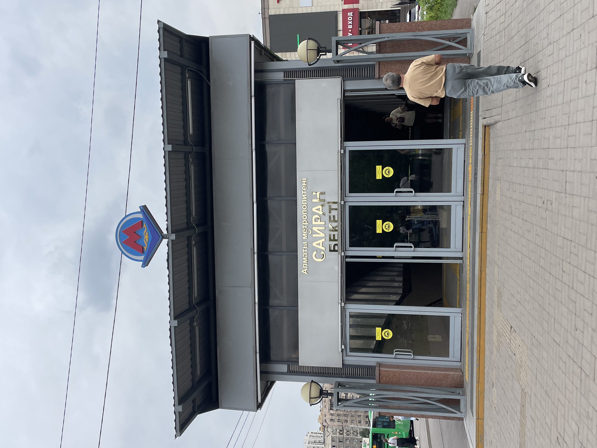 The image shows the entrance to the "Sairan" metro station in Almaty, Kazakhstan. The station entrance is a modern, gray structure with a large, blue and red metro logo above the entrance. The words "Алматы метрополитені" and "САЙРАН БЕКЕТІ" are written in gold letters above the glass doors. Two people are entering the station, and another person is walking away from the entrance on a paved sidewalk. A green bus is visible in the background, as well as some buildings and a cloudy sky. The scene appears to be a typical day in a city.