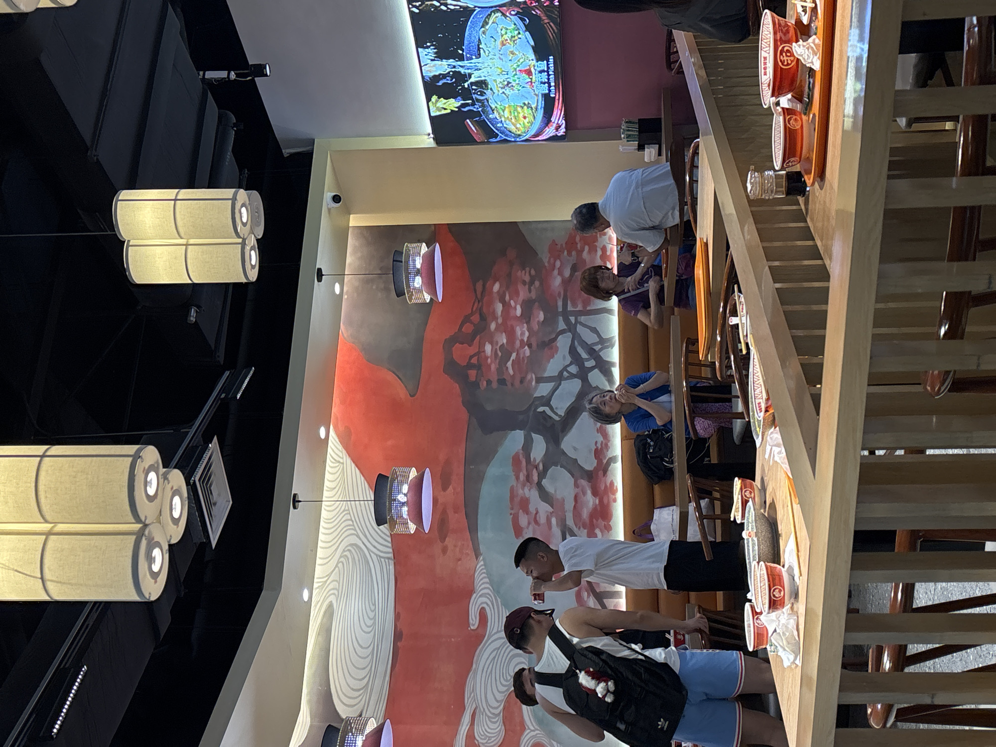 The image depicts the interior of a restaurant, likely located in Jianyang City, China, given the context. The scene is set in a dining area with wooden tables and chairs. The walls feature a large mural with a red and white abstract design, suggestive of clouds and a tree with red blossoms. Overhead, multiple pendant lamps with cylindrical white shades illuminate the space.
In the foreground, a wooden table displays several bowls and trays with food, indicating recent or ongoing dining. Two young men are standing near the table, one holding a drink. Further back, an older woman and a man are seated at a table, engaged in conversation. On the right side of the image, a large flat-screen television displays a close-up of a steaming pot of food. The overall atmosphere appears casual and communal, typical of a restaurant setting.