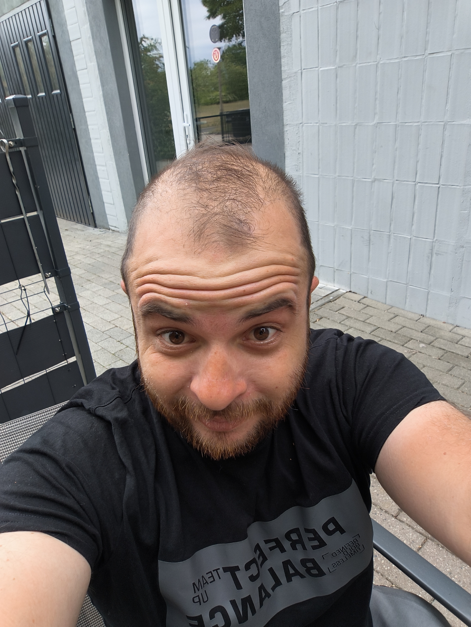 A selfie taken outdoors shows a white male with a reddish-brown beard and thinning hair. The man is wearing a black t-shirt with gray lettering. He is seated in a dark-colored chair. The background includes a white wall with a rectangular pattern and a dark metal fence. Paved brick is visible on the ground. A glass door with a thin white frame and an opaque sticker is behind the subject. The location is in the city of Unknown, Unknown.